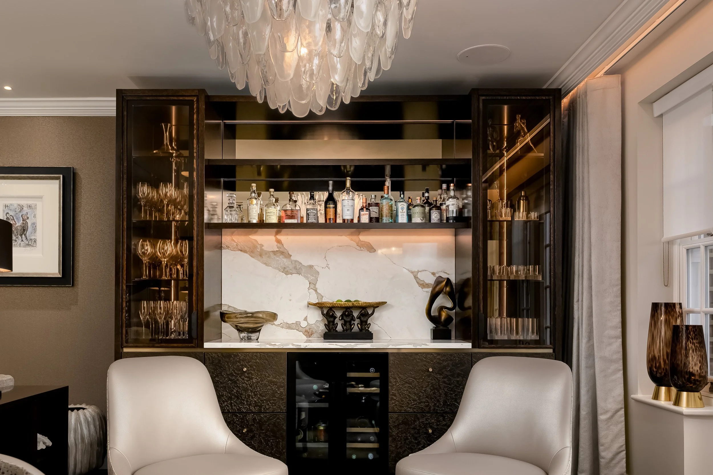 Interior view of a living room featuring a bar area with bottles of alcohol on a shelf, a marble backsplash, decorative statues, and a chandelier with glass drops hanging from the ceiling. There are white chairs in the foreground and a window with curtains on the side.