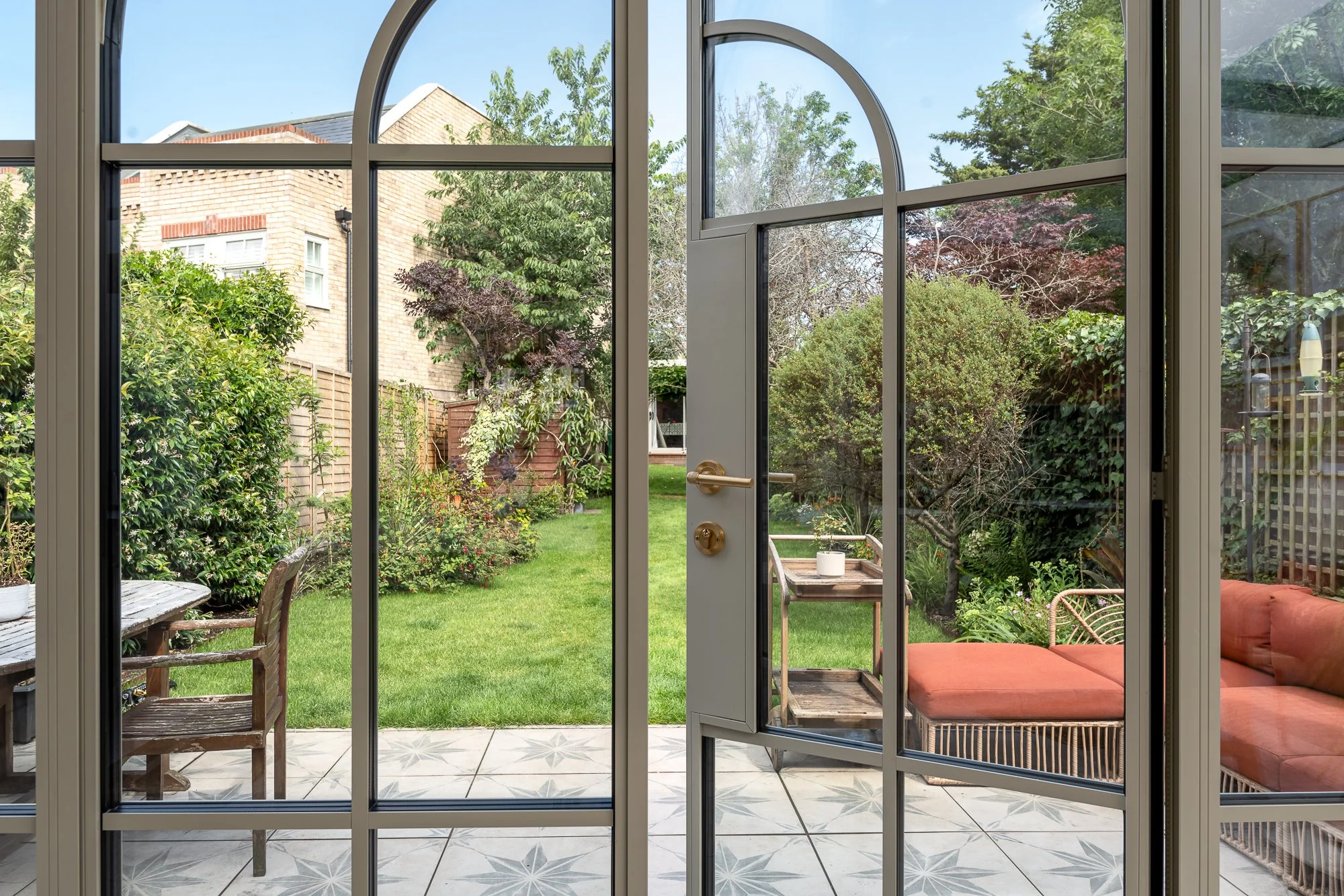 An interior view through arched glazed garden doors opening onto a patio and landscaped garden. The image highlights the slim metal frames, curved glazing detail and the connection between the indoor living space and outdoor area.