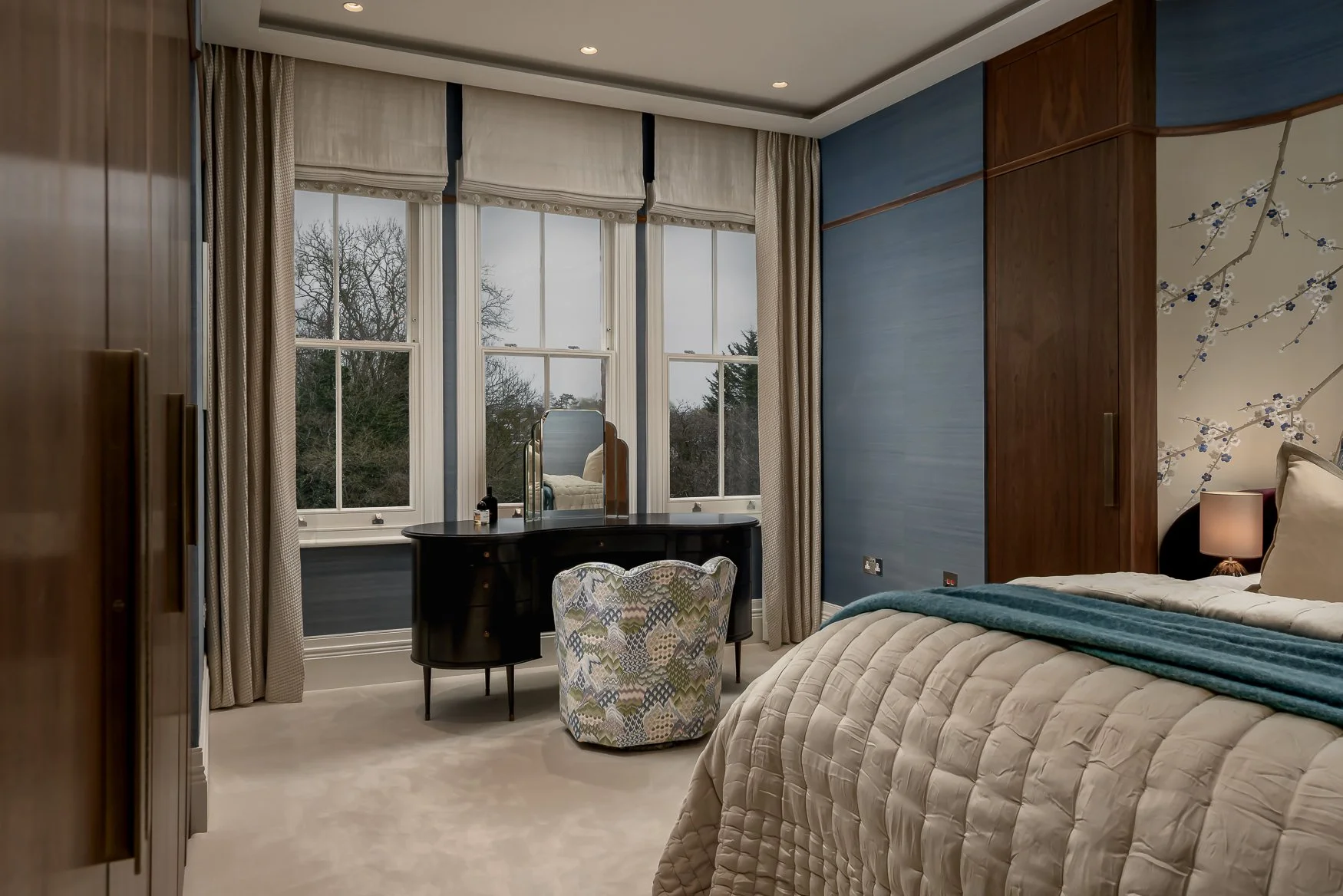 A beautifully decorated bedroom with a large window, a vanity table, a patterned armchair, a bed with beige quilted bedding, a bedside lamp, and a blue accent wall with floral mural.