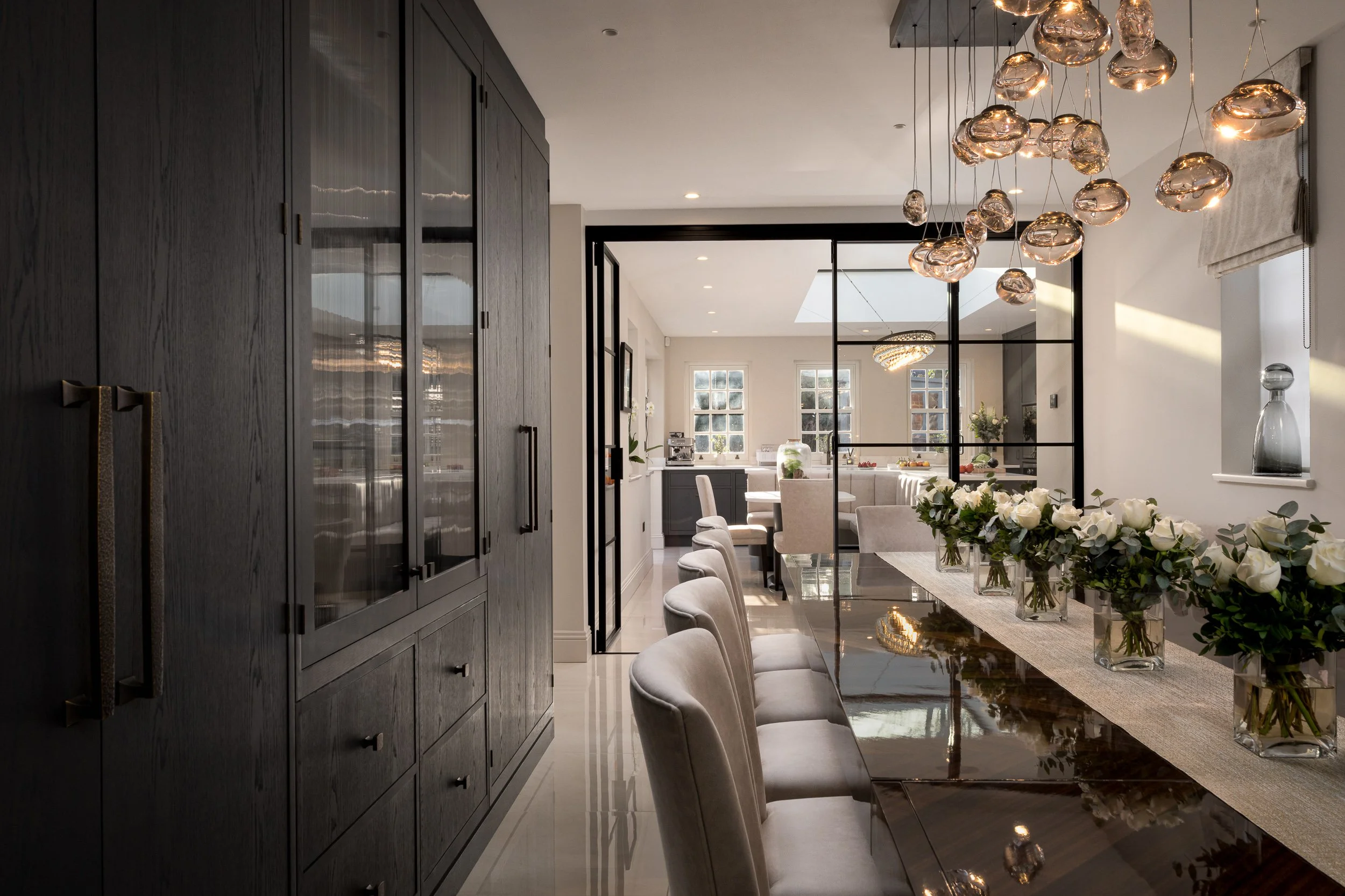 Luxury dining room with dark cabinetry, glass partition doors, sculptural pendant lighting and floral table styling.