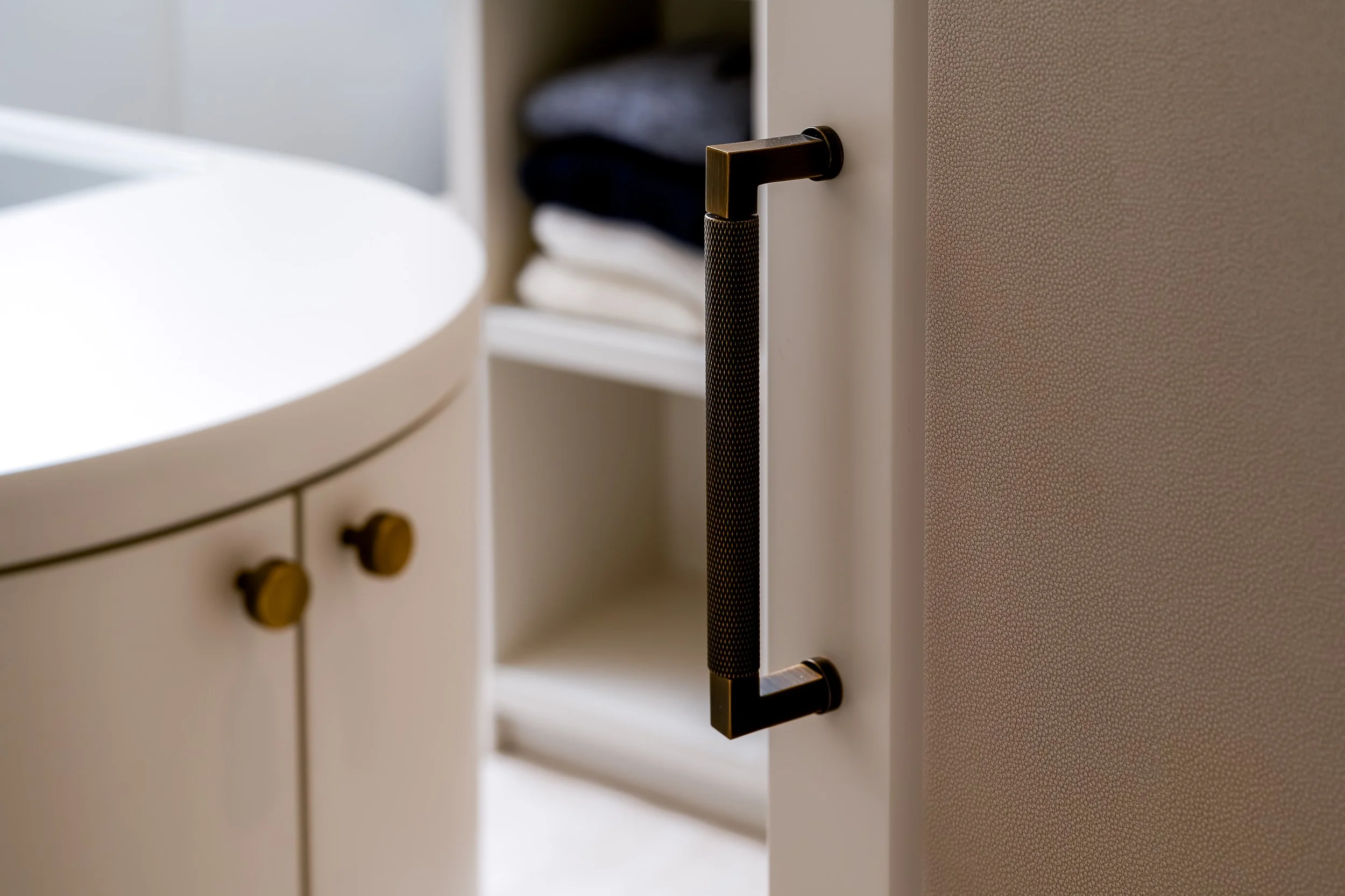 Close-up of a black brass handle on a white cabinet door with a textured beige wall beside it, and a blurred open cabinet with folded clothes inside.
