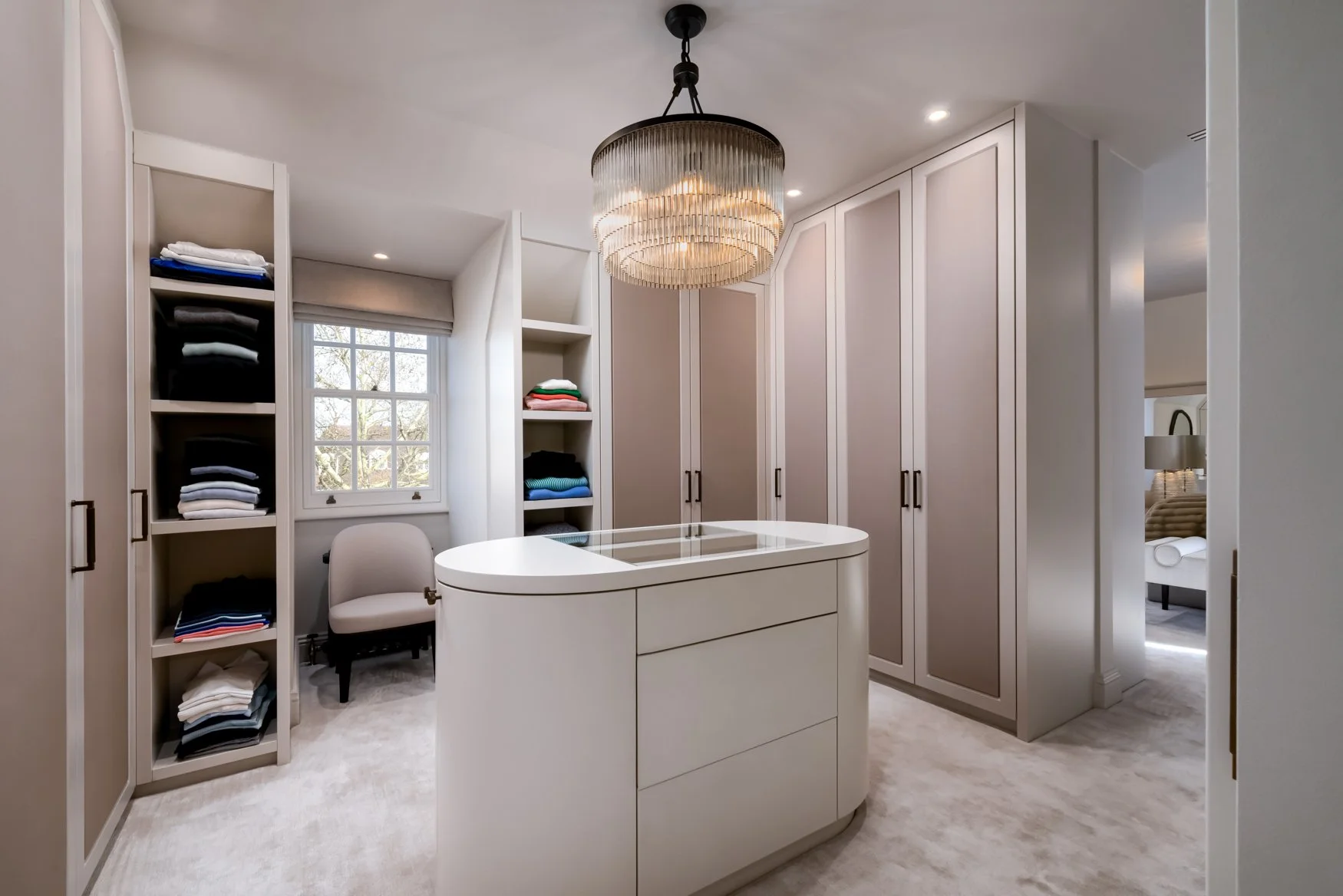A walk-in closet with built-in shelves filled with folded clothes, a small window, a white chair, and a modern chandelier hanging from the ceiling.
