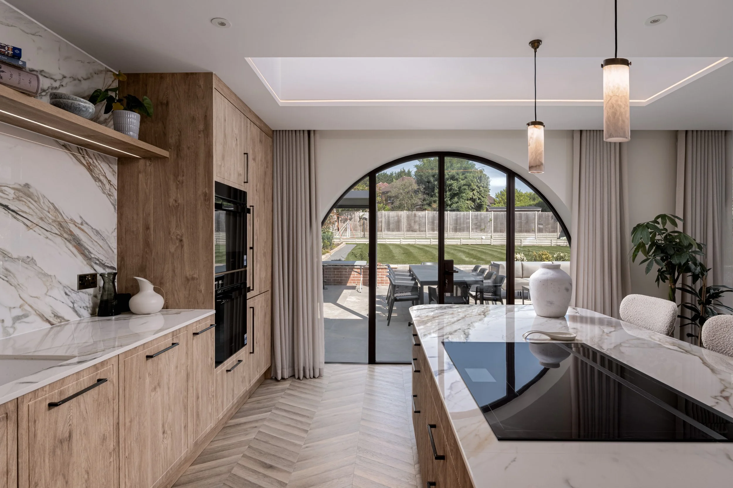 A modern kitchen interior with an arched glazed door opening onto an outdoor patio and garden. The image highlights the curved glazing, dark frame, natural light and how the door connects the kitchen with the outside space.