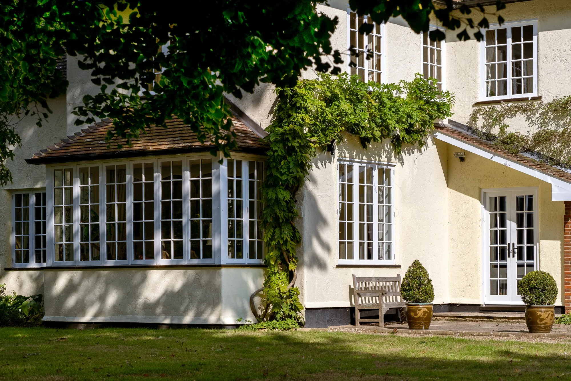 An exterior view of a traditional house with white timber-style windows and French doors surrounded by mature greenery. The image highlights the classic window proportions, multiple glazed panels and how the doors and windows shape the character of t