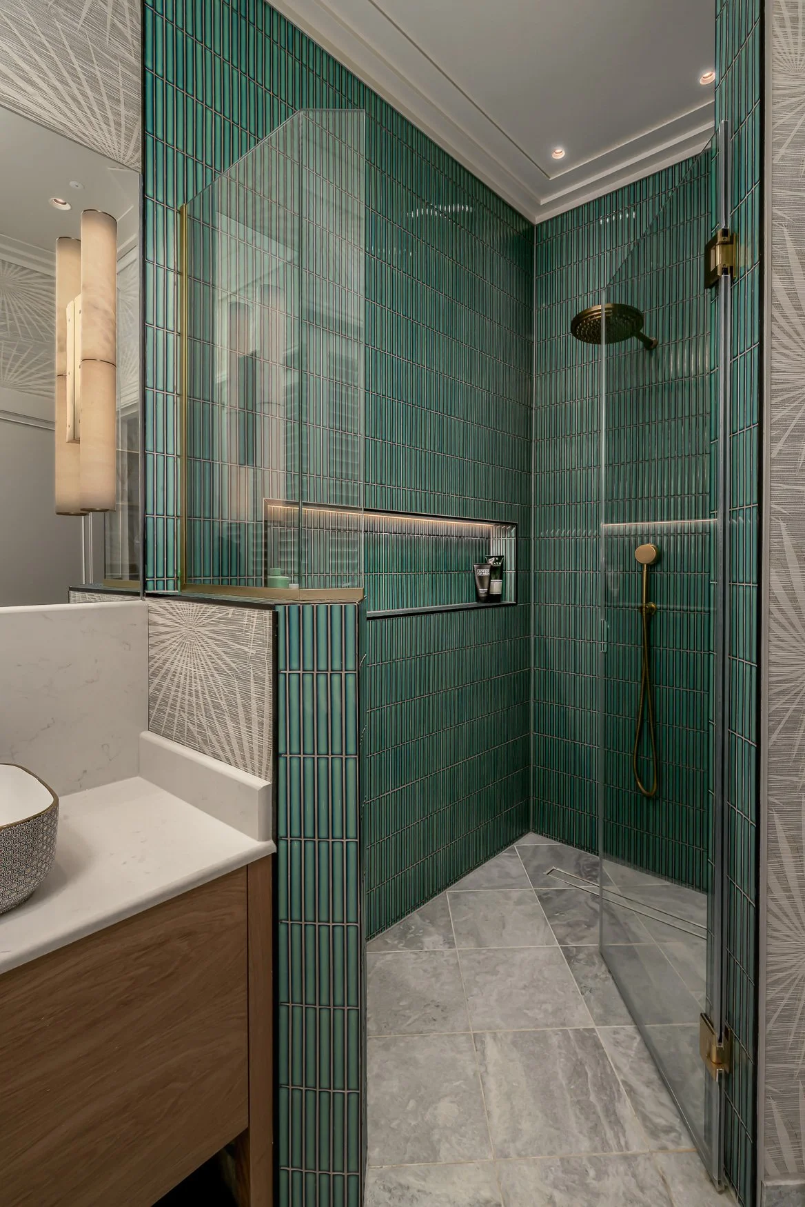 Modern bathroom with green tiled walk-in shower, gold fixtures, marble flooring, and a partial view of a vanity with a white marble top and wooden cabinet.