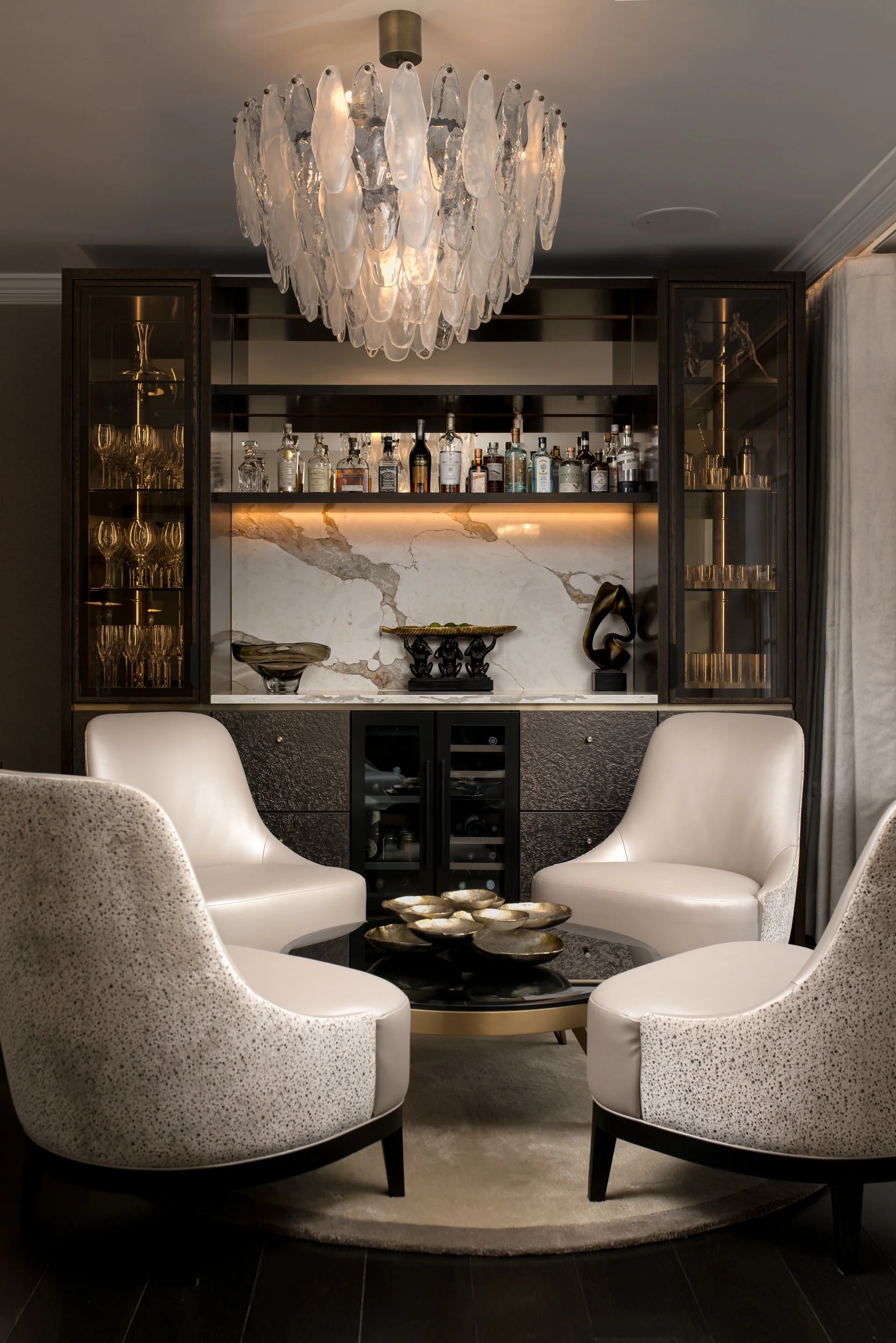 A modern living room with four white armchairs surrounding a black coffee table on a round beige rug. Behind the seating area, there is a bar with various bottles and glassware, a marble backsplash, and decorative objects. A large chandelier made of 