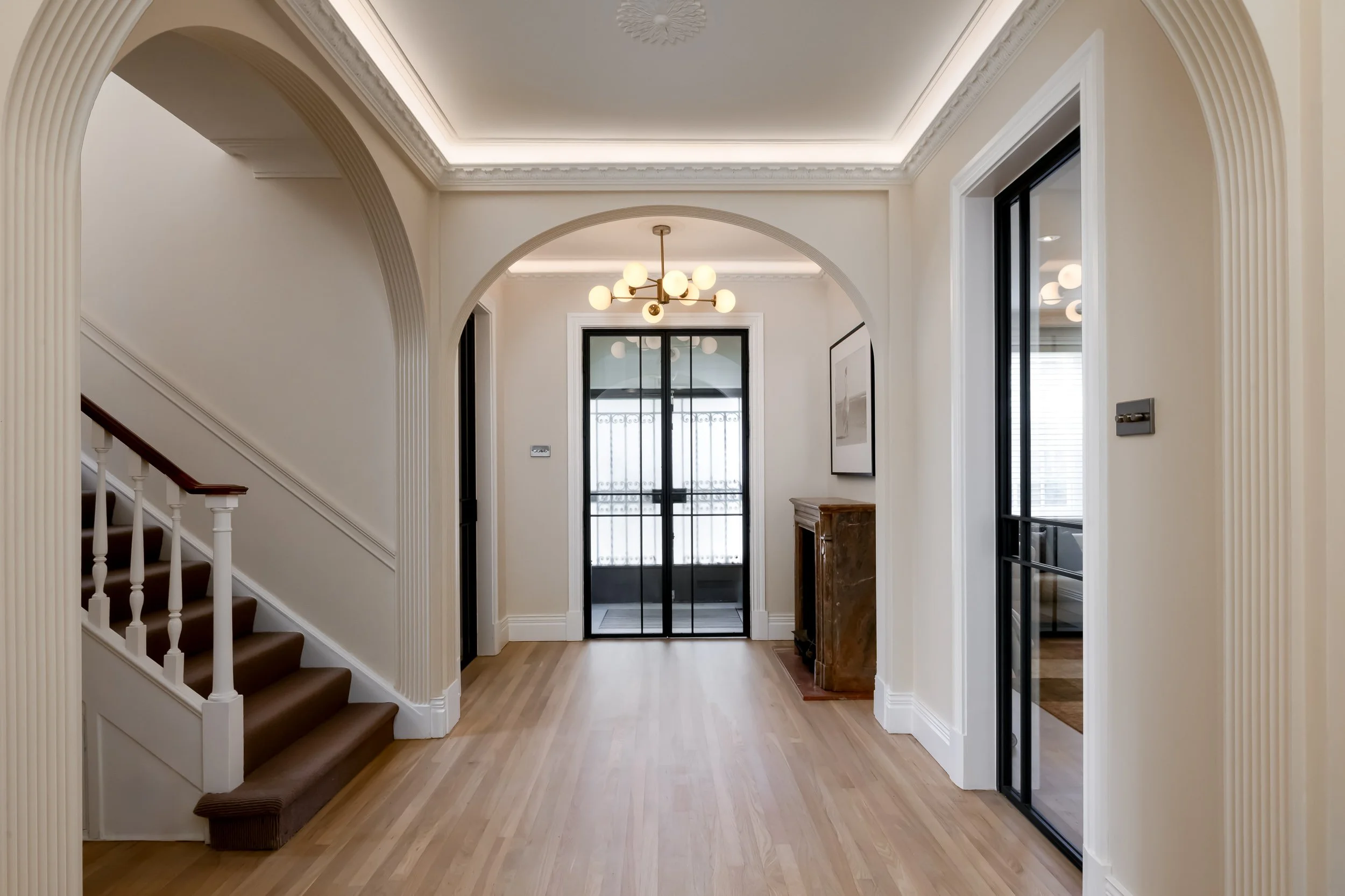 Bright entrance hall in a renovated London home with arched openings, staircase and black-framed glazed doors. Home in Hampstead, London.