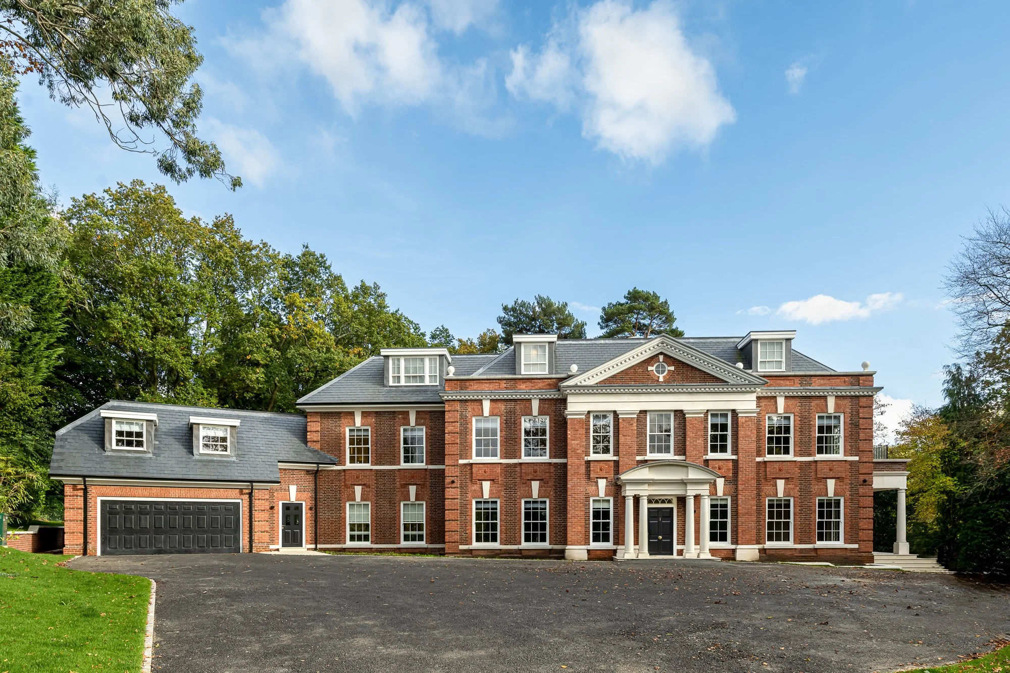 Front elevation architectural photograph of a grand, luxury Neo-Georgian style residential mansion. The exterior features symmetrical red brickwork, classic white stone columns framing a central entrance portico, traditional sash windows, and a prist