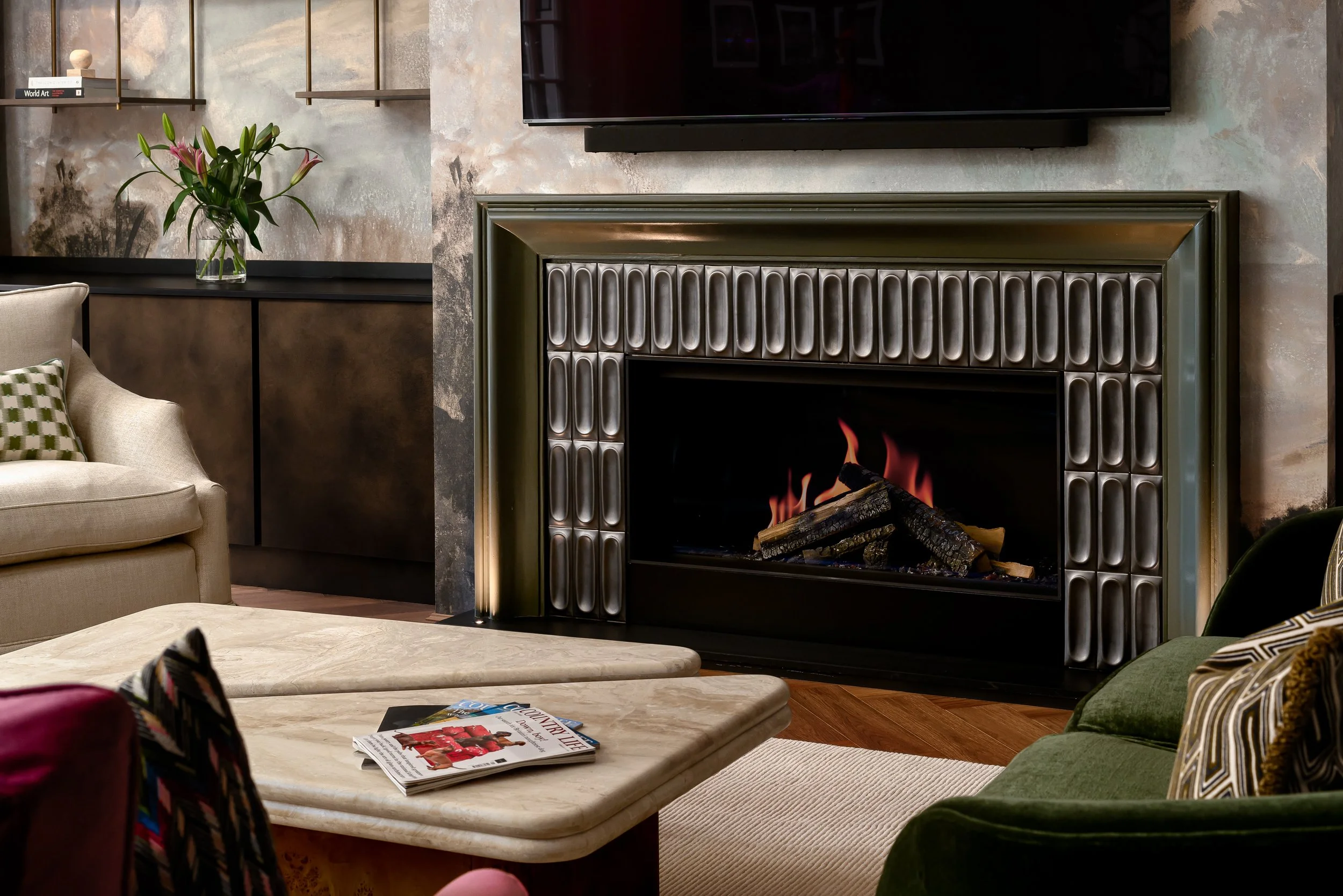 A luxury living room fireplace detail photographed in London, featuring a sculptural tiled fireplace surround, marble coffee table, layered seating, textured wall finishes, bespoke shelving, flowers and warm firelight. The image focuses on material q