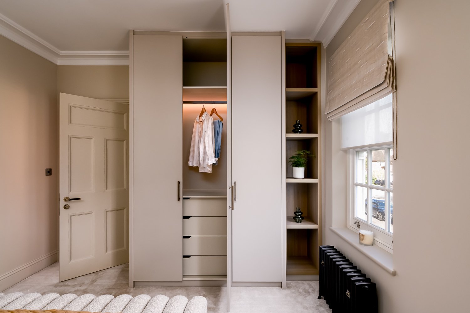 A bedroom with a beige door, a built-in wardrobe with open and closed sections, and a window with beige curtains. The wardrobe has hanging clothes, shelves with decorative items and a potted plant. There is a black radiator below the window and a sma
