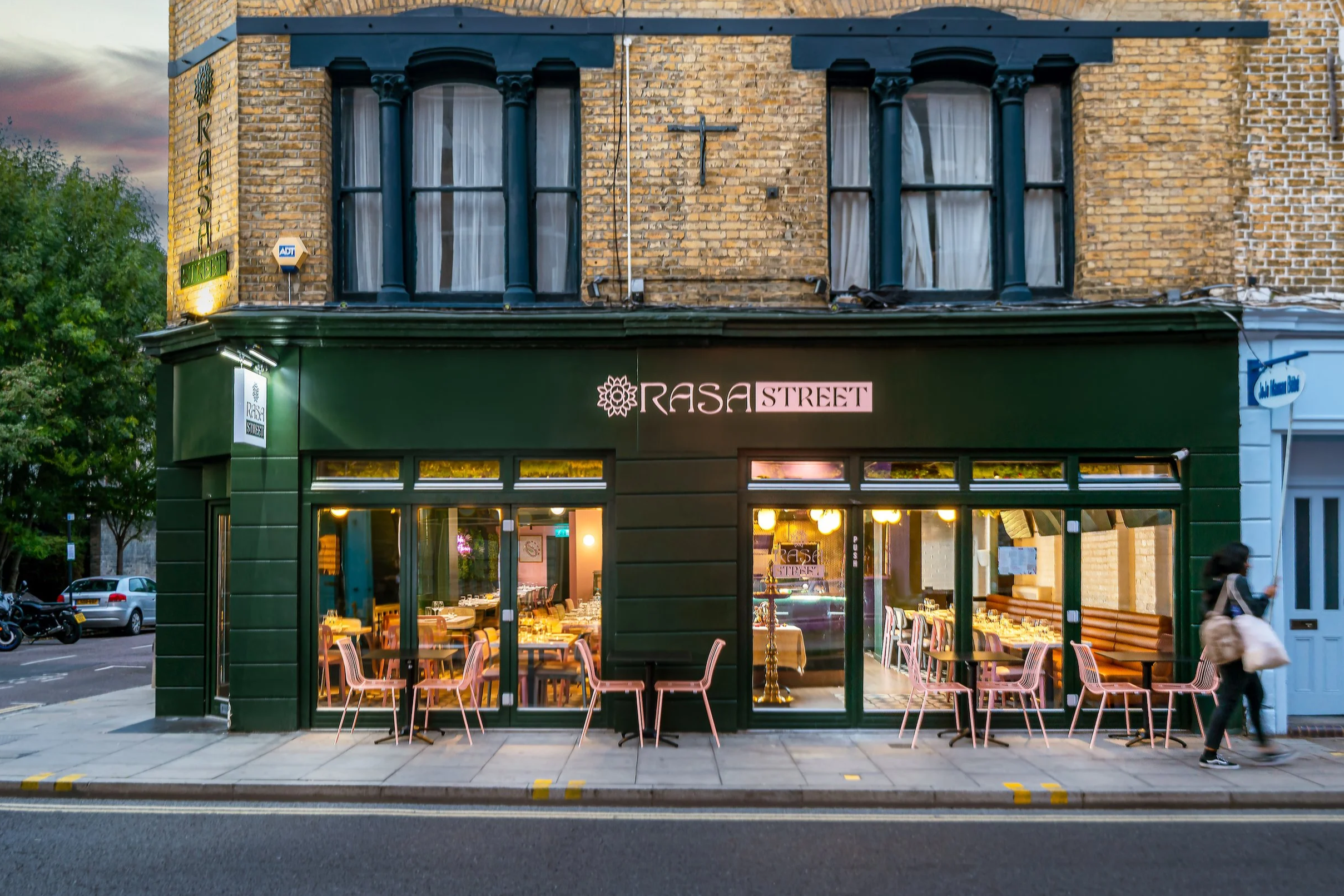 Exterior of a restaurant named Rasa Street with large glass windows, pink chairs outside, and a brick upper facade with black window trims. Some trees and parked cars are visible nearby. Hackney restaurant, London.