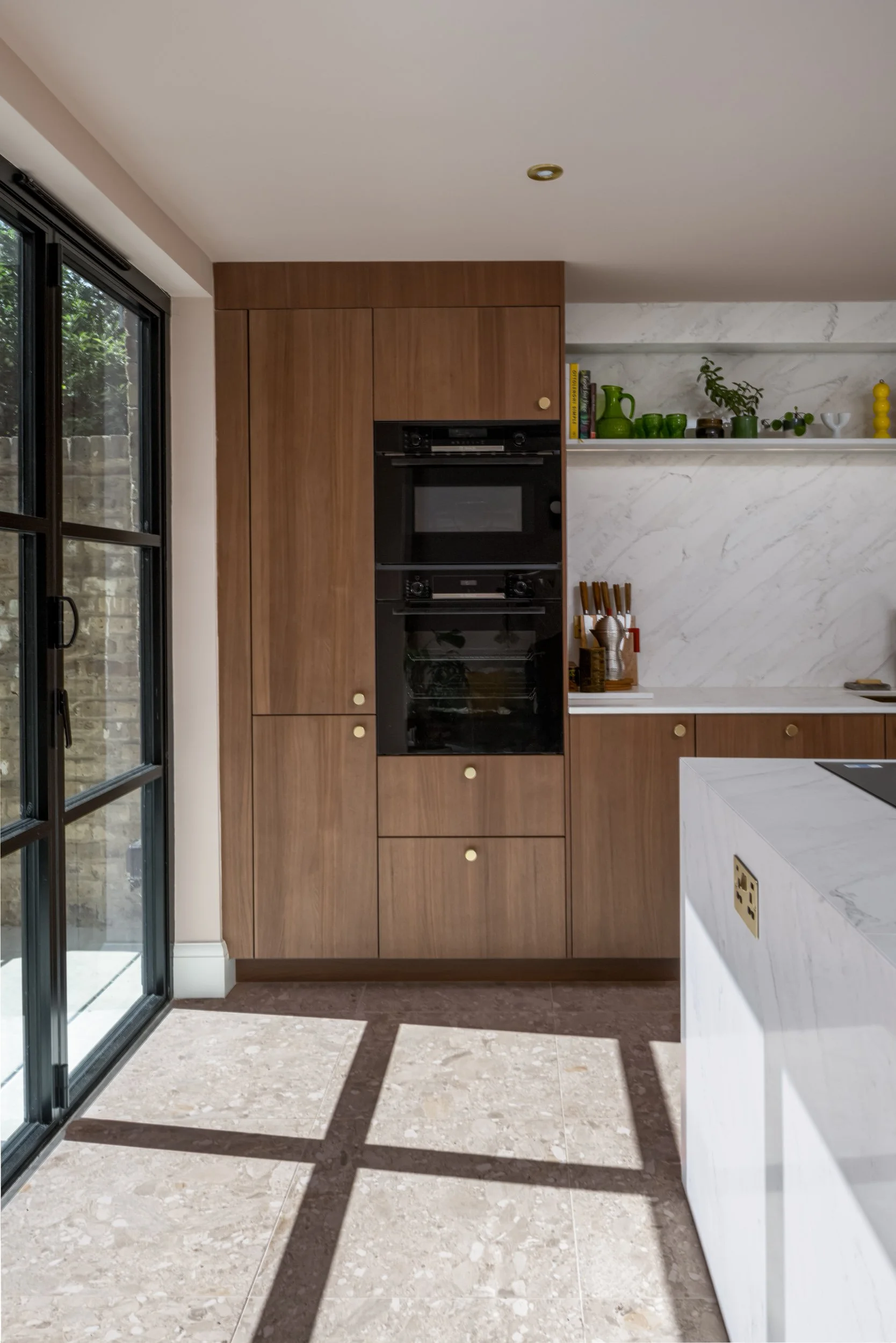 Modern kitchen with wooden cabinets, marble backsplash, and a white marble island, with sunlight casting shadows on the beige tiled floor.