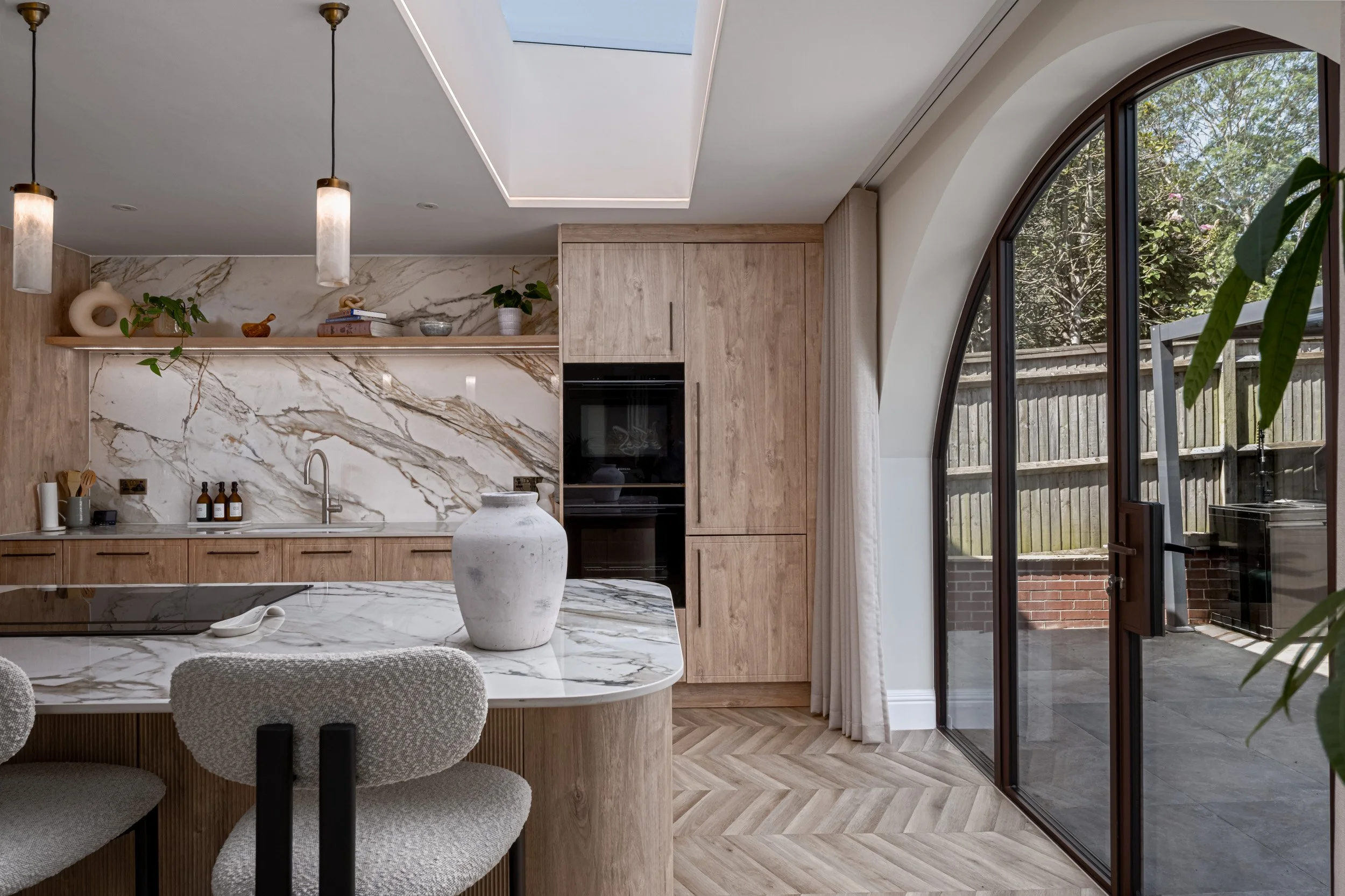 A detailed view of a kitchen interior with an arched glazed door opening onto an outdoor patio. The image highlights the curved glazing, dark frame, natural light and how the door connects the kitchen with the outside space.