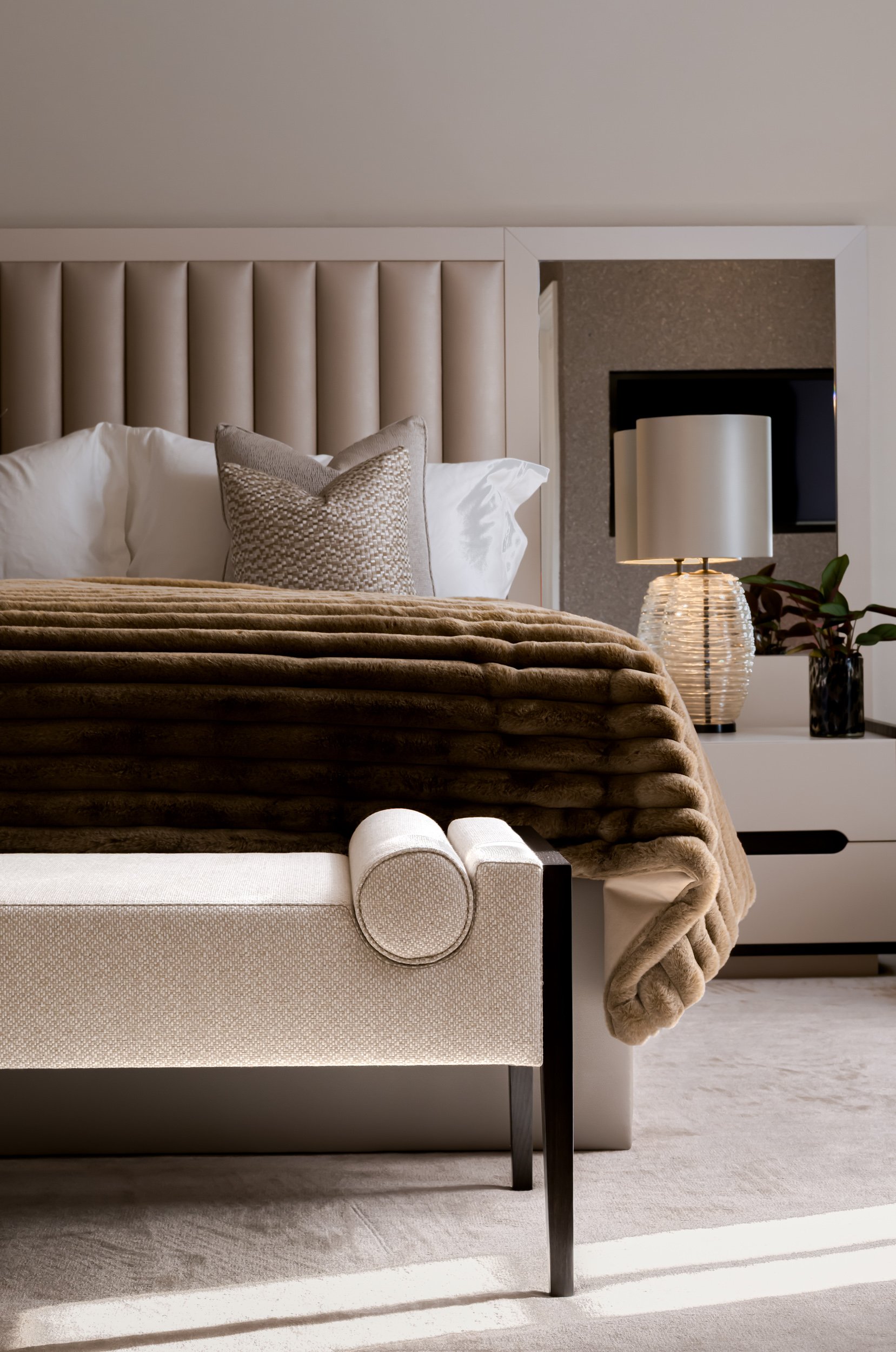 A cozy bedroom with a beige upholstered headboard, white pillows, a textured throw pillow, and a blanket. A cream-colored bench with rounded armrests is at the foot of the bed. To the right, a nightstand with a textured glass lamp and a potted plant 
