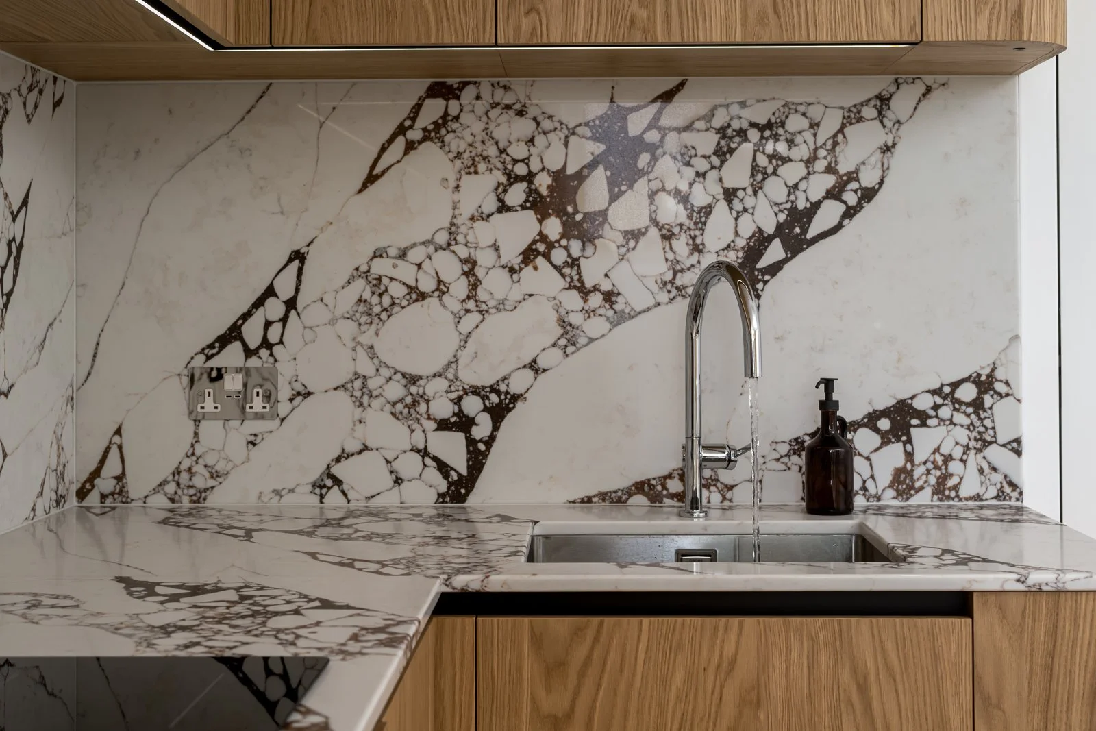 Modern kitchen with marble countertops and backsplash featuring white and dark brown veining, a stainless steel sink with water running, a brown soap dispenser, and wooden cabinetry.
