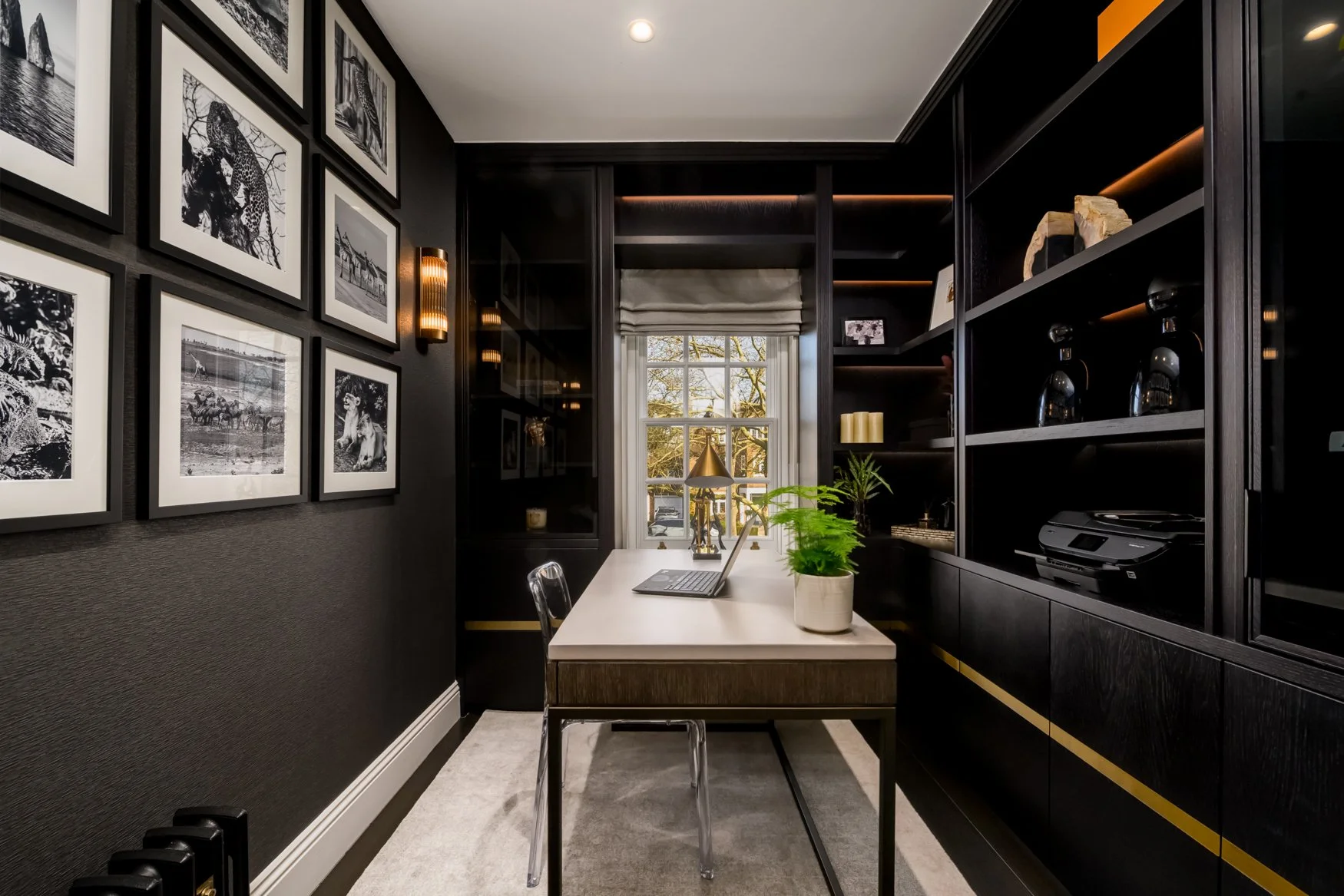 A home office with black walls and neat decor, featuring a white desk with a laptop and a potted plant, artworks of animals on the wall, a window with a view of trees outside, and built-in black bookshelves with decorative items.