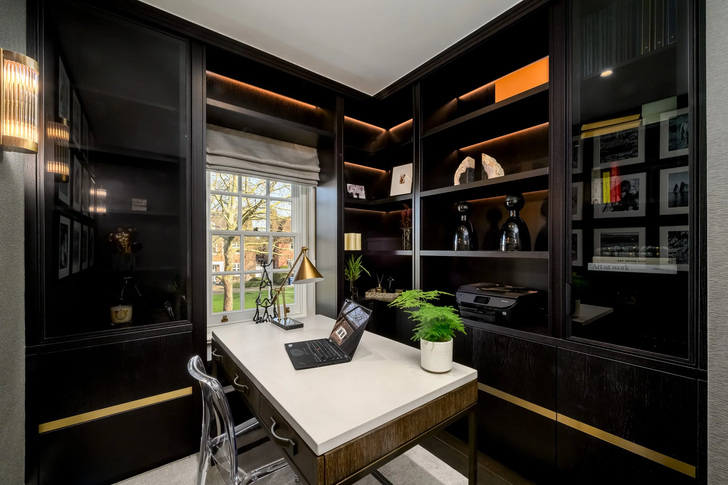 A home office with black built-in shelves and cabinets, a white desk with a laptop, a potted fern, decorative items including sculptures and books, and a window with a view of trees outside.