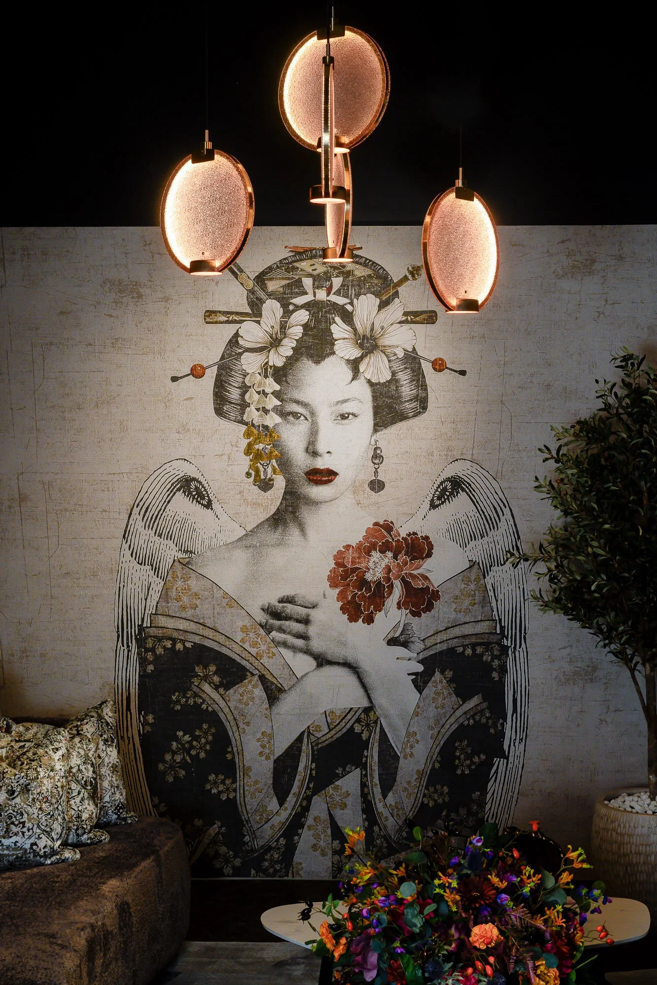 A floral arrangement with vibrant multicolored flowers and greenery on a white table, in front of a large wall mural of a geisha with traditional hair accessories and makeup, beneath three hanging light fixtures. Wallpaper detail shot from showroom