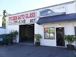 The image shows the exterior of Golden Auto Glass, an auto glass repair shop, with a sign displaying the business name and phone number. There is a parking area and indoor seating area.
