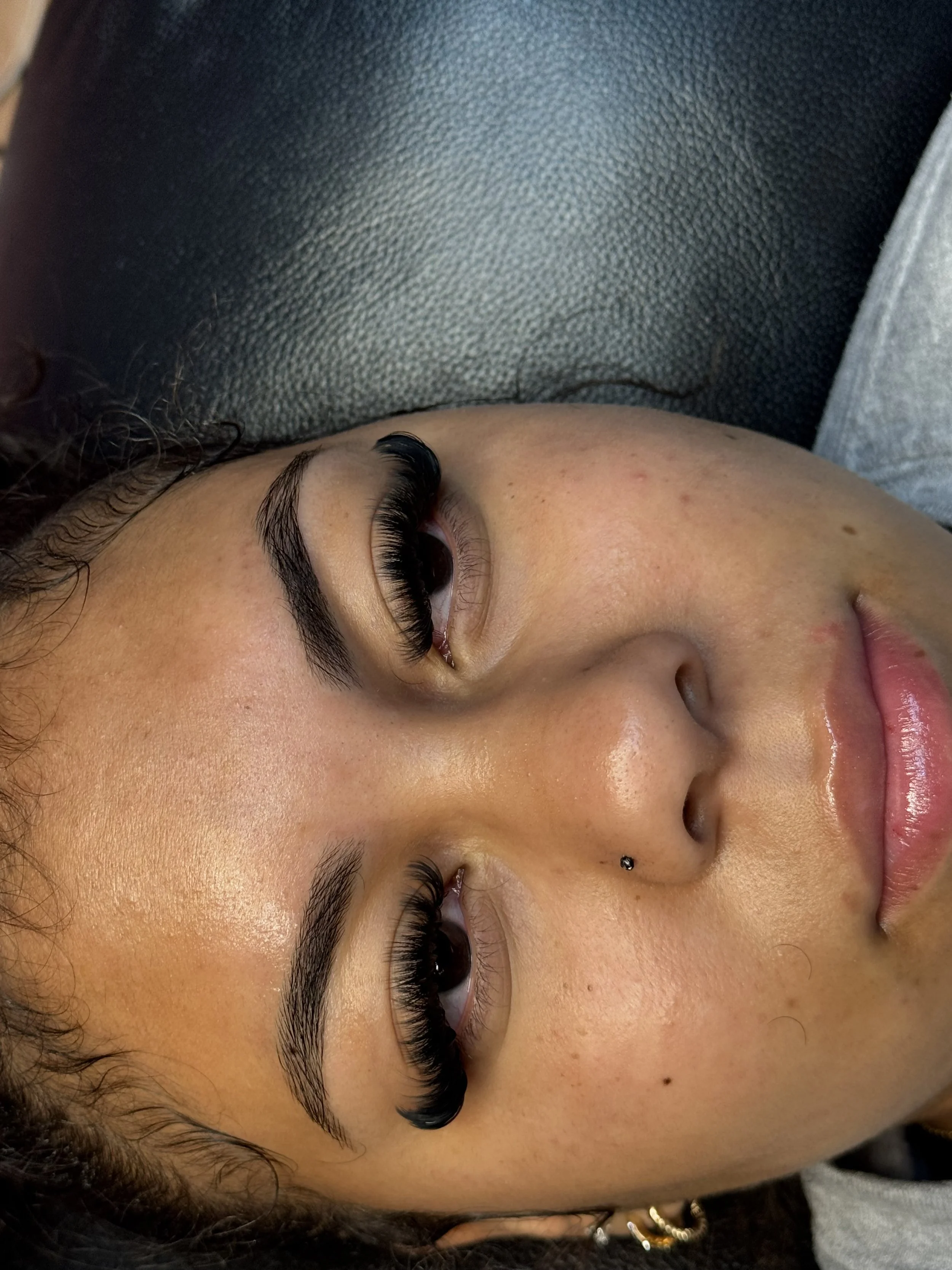 Close-up of a woman lying on a black surface, showing her face with makeup, long eyelashes, and a nose piercing.