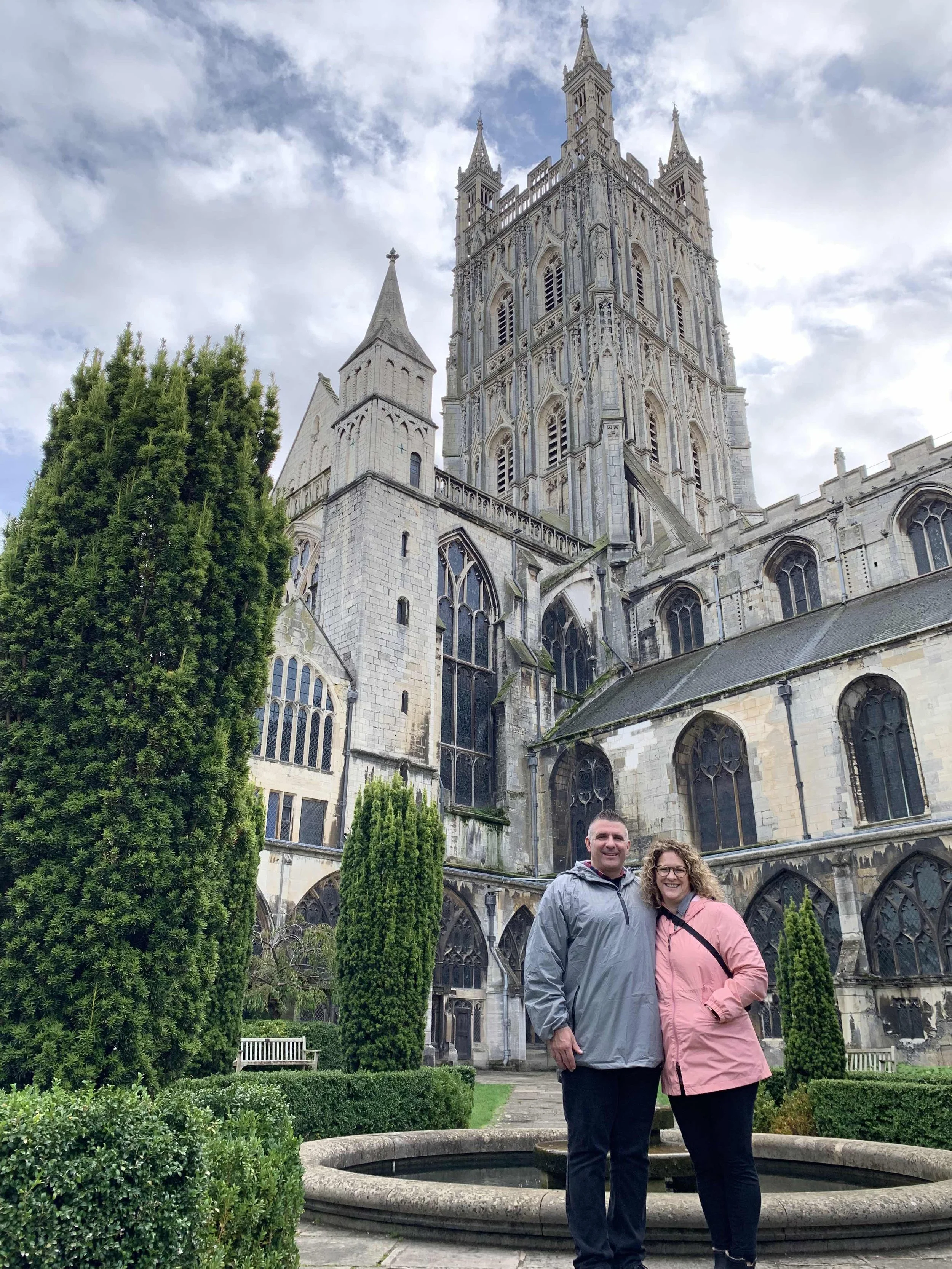 A couple standing in front of an ancient cathedral with a tall spire, surrounded by well-trimmed bushes and trees, under a cloudy sky.