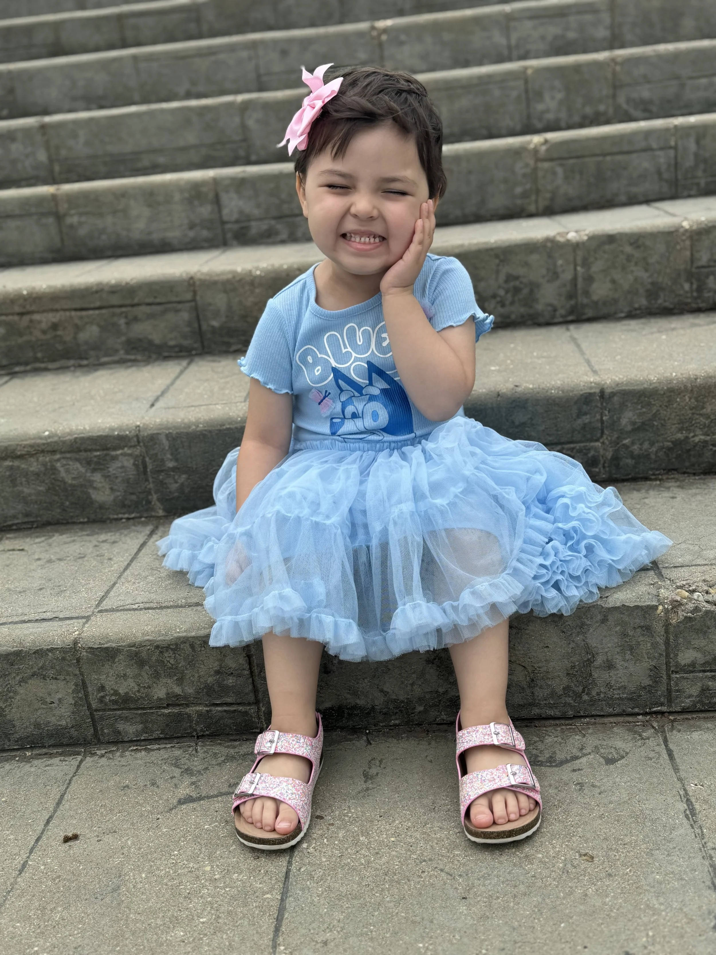 A young girl with short dark hair, wearing a blue t-shirt with a cartoon character and the words 'Blue' on it, a fluffy blue tutu, pink glittery sandals, and a pink bow headband, sitting on stone steps outside, touching her face and smiling.