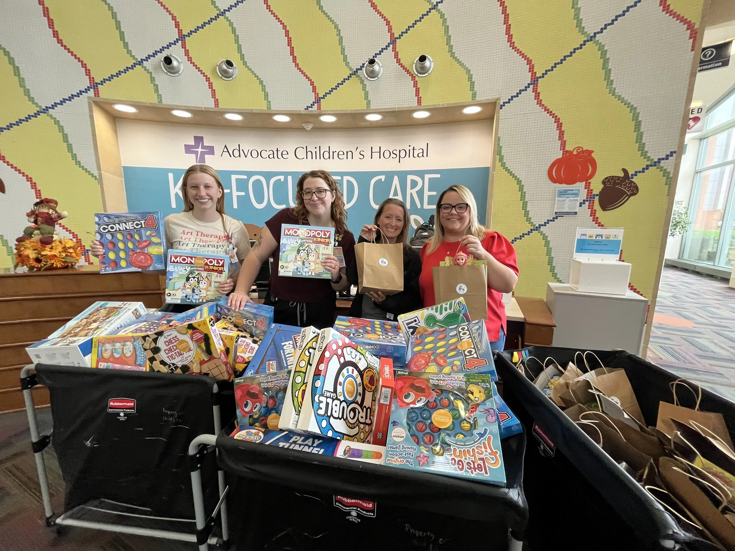 Four women standing behind a table full of toys at a charity event for Advocate Children's Hospital, smiling and holding children’s books and gift bags, with a backdrop that reads 'KAISERED CARE'.