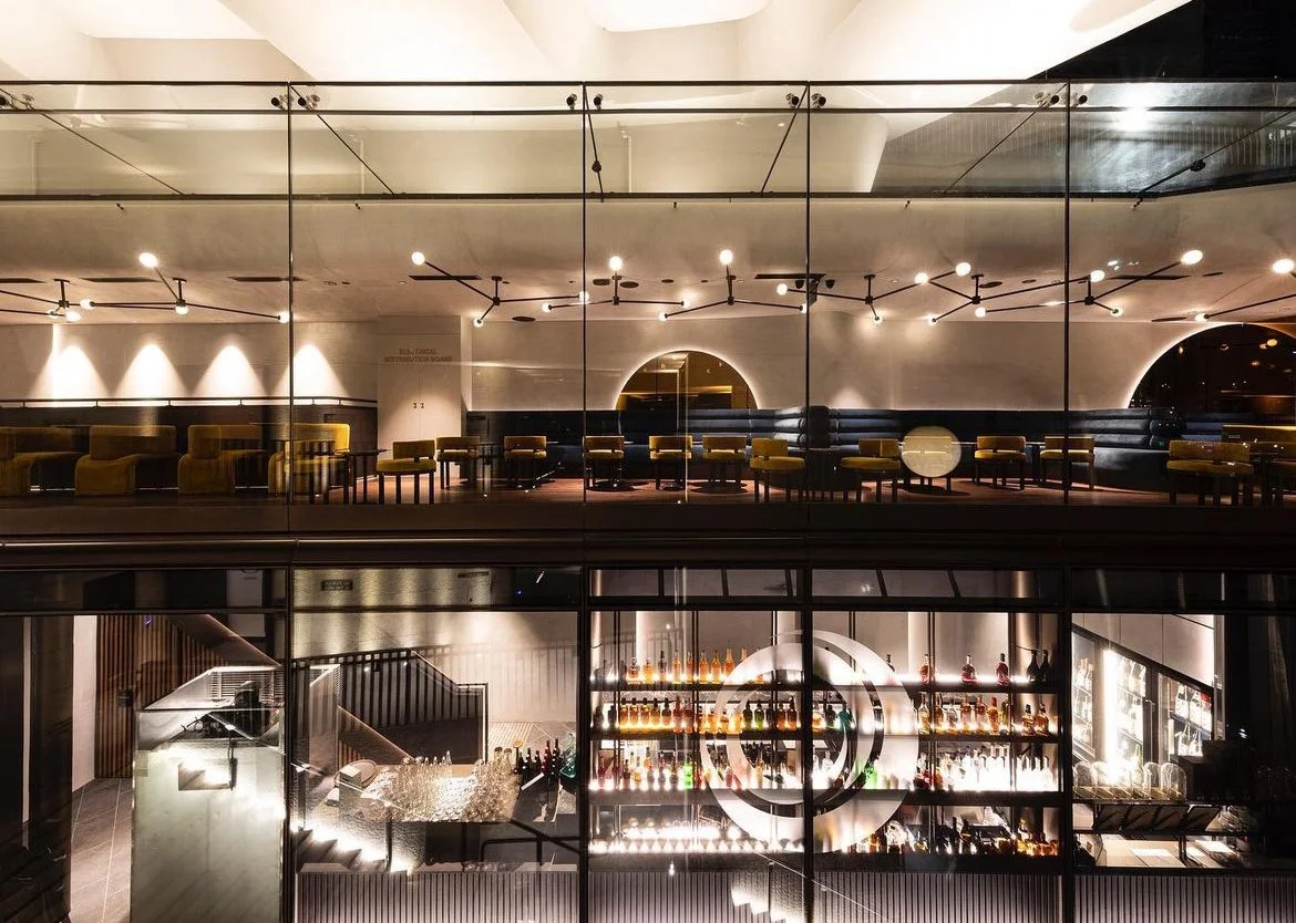 Interior of an upscale bar and lounge area seen through large glass windows, featuring a well-lit bar with bottles of liquor, and stylish seating with yellow chairs and modern lighting fixtures.