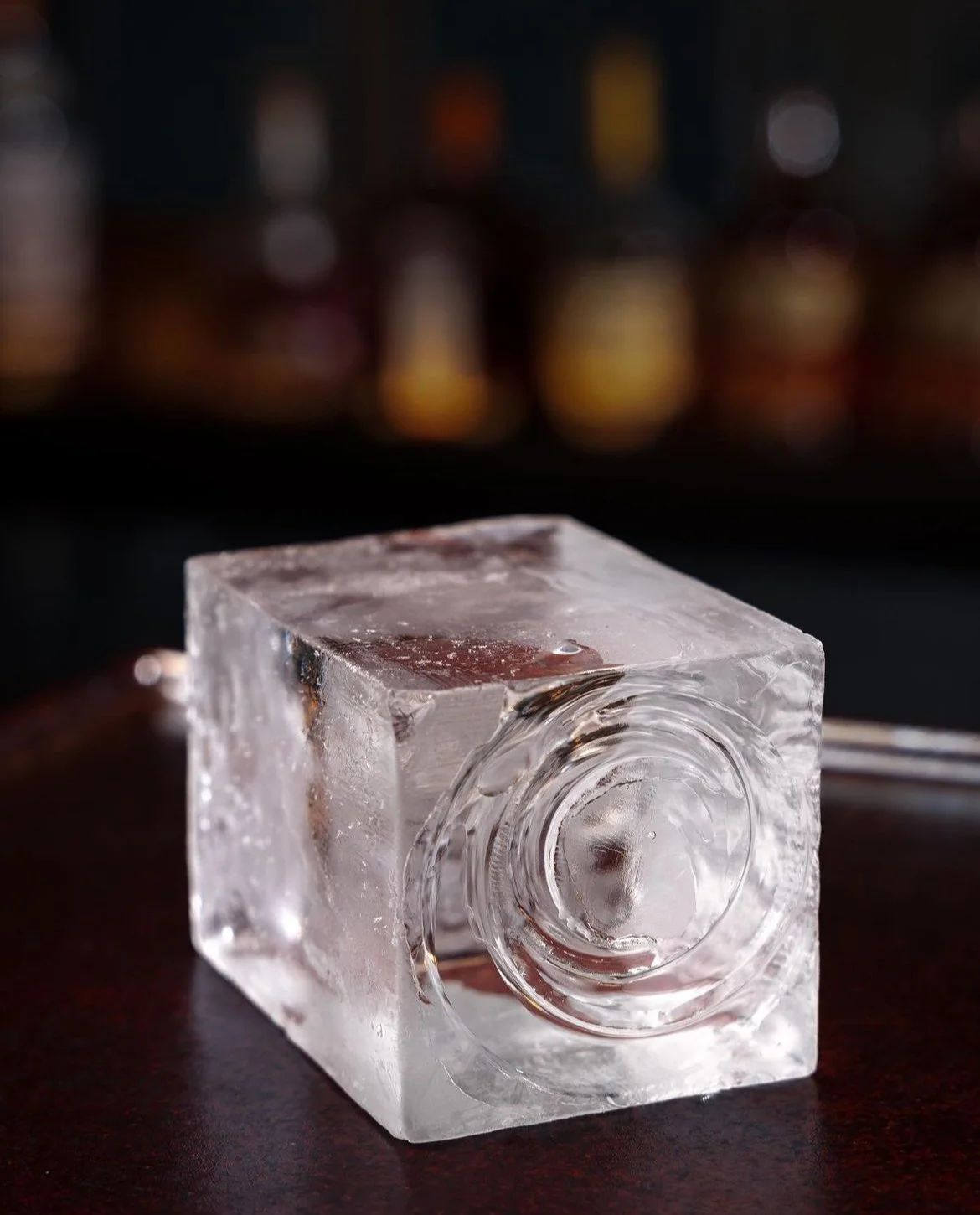 Close-up of a clear ice cube on a dark surface with a blurred bar background.