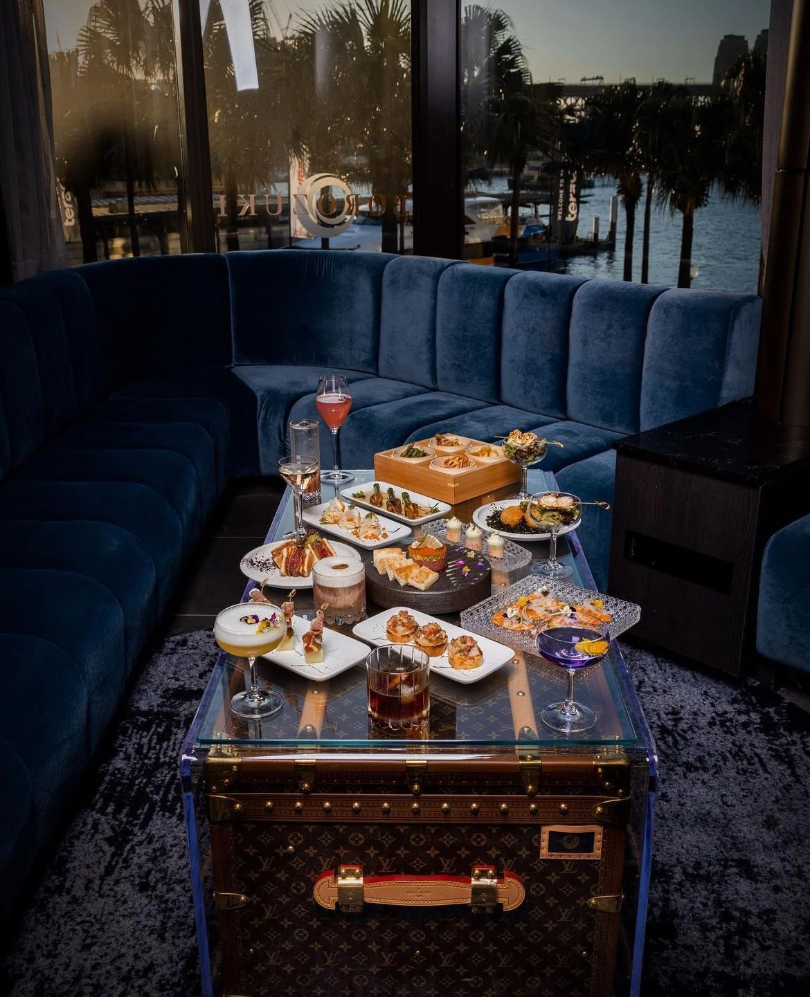A luxurious room with a blue velvet sectional sofa behind a glass table filled with various gourmet appetizers and cocktails. Large windows overlook a marina with boats and palm trees.