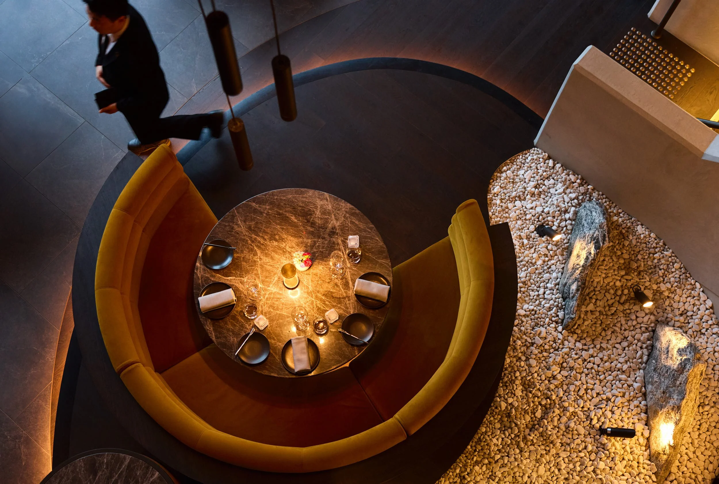 Overhead view of a round marble table set for six with black plates, napkins, glasses, and cutlery. The table is surrounded by a curved mustard yellow banquette. To the right, a stone and pebble interior feature with large rocks and small spotlights. A person in dark clothing is walking on a curved dark wooden floor at the top left of the image.