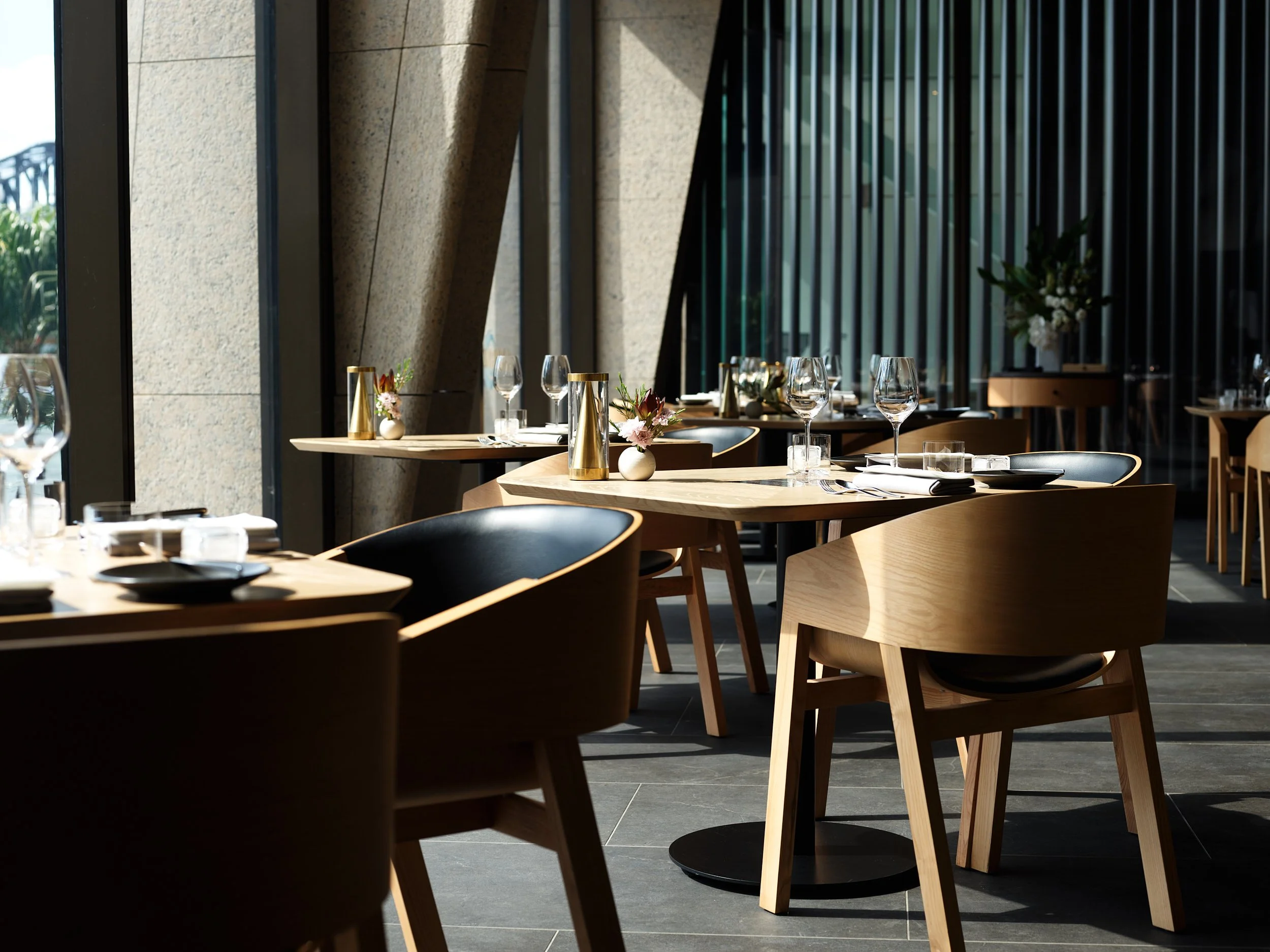 Empty dining tables set with wine glasses, small vases with flowers, and gold-colored bottles in a modern restaurant with large windows and wooden chairs.