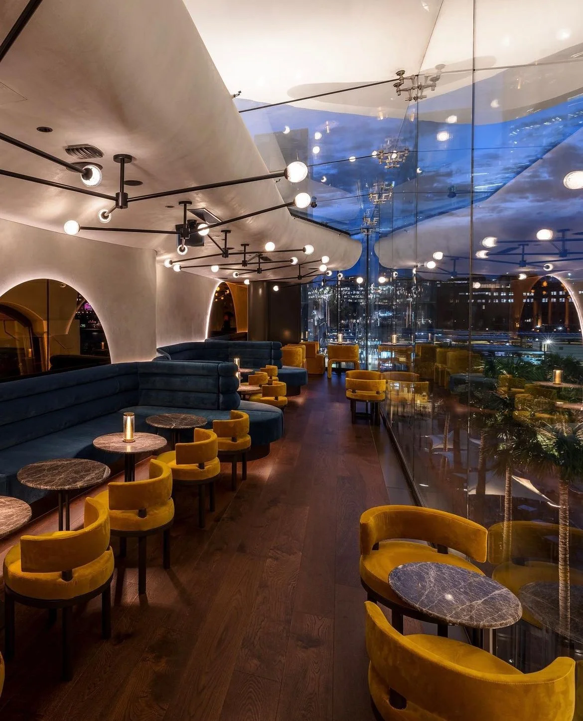 Interior of a modern restaurant or lounge with yellow velvet chairs, marble-top tables, dark wood flooring, and innovative ceiling lighting fixtures. Large glass windows reveal a cityscape at night.