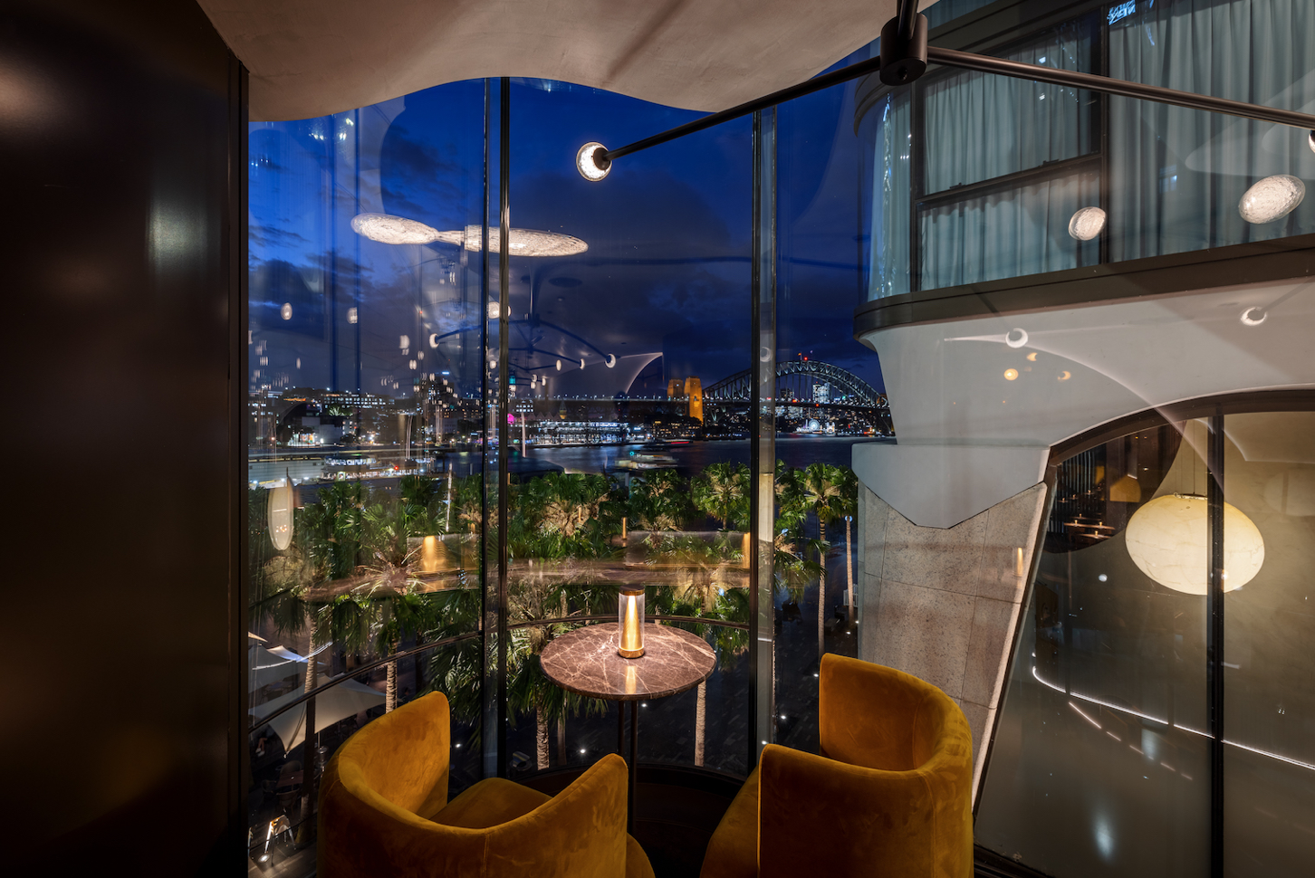 Interior view of Oborozuki’s bar at night with large glass windows, yellow velvet chairs, small round tables with a lamp, and a cityscape with a bridge visible outside.