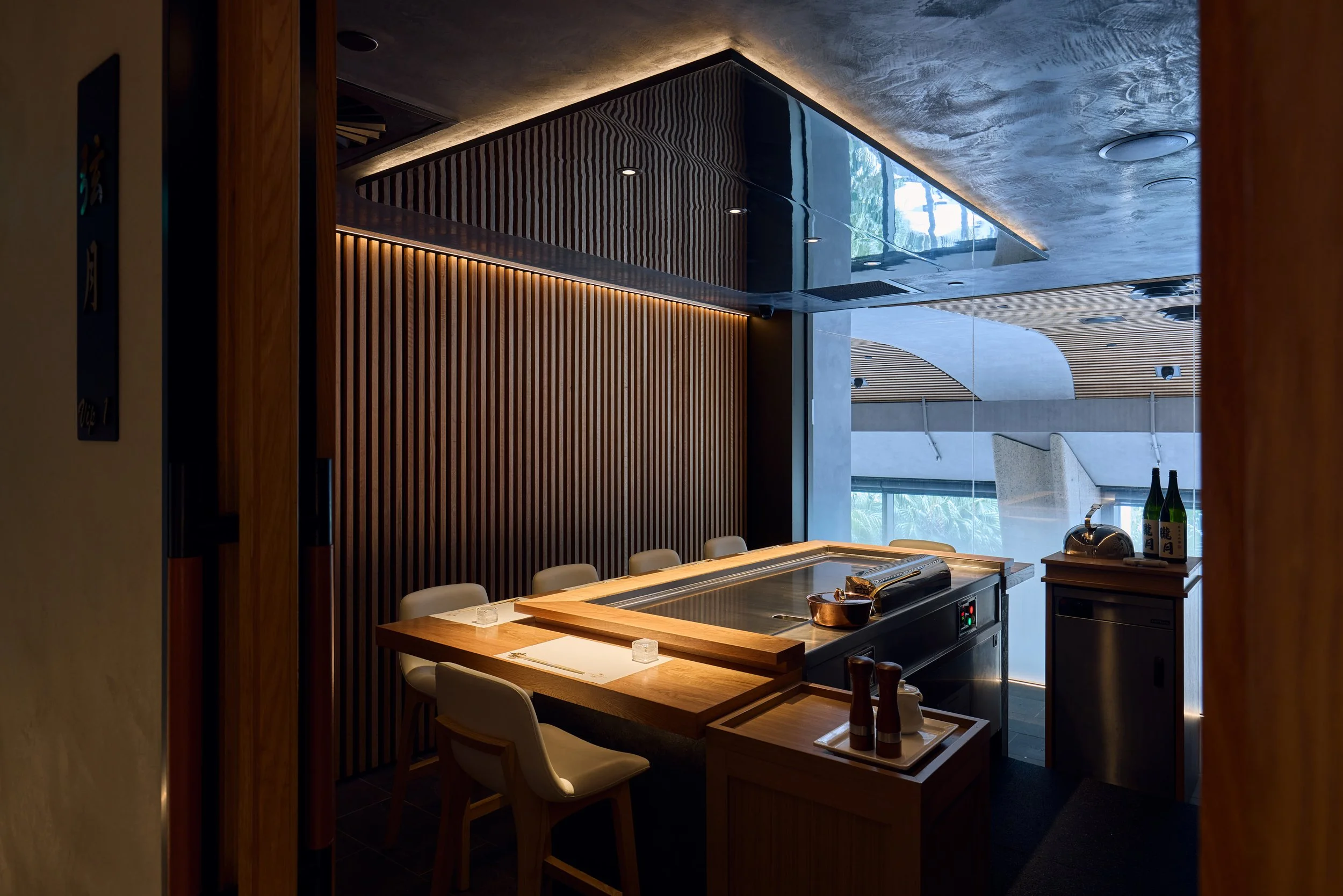Interior of a modern Japanese restaurant with a wooden counter and seating, a teppanyaki grill, bottles of sake, and a view of large windows with natural light.