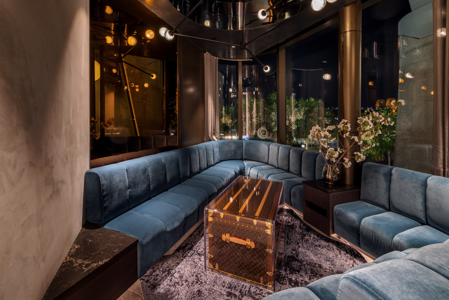 Luxurious seating area with blue velvet sectional sofa, modern lighting fixtures, large windows with curtains, a glass coffee table Louis Vuitton suitcase in a stylish contemporary lounge at night.