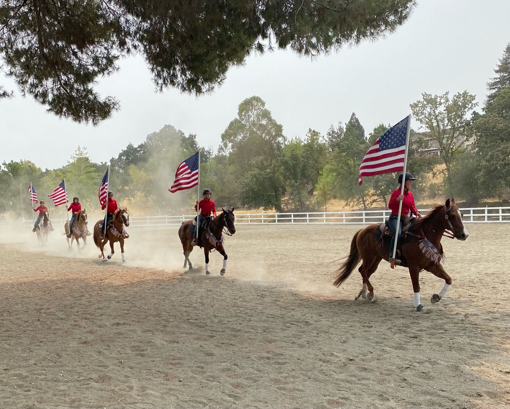 People riding horses in a rodeo or parade, carrying American flags, outdoors with trees and fencing in the background.