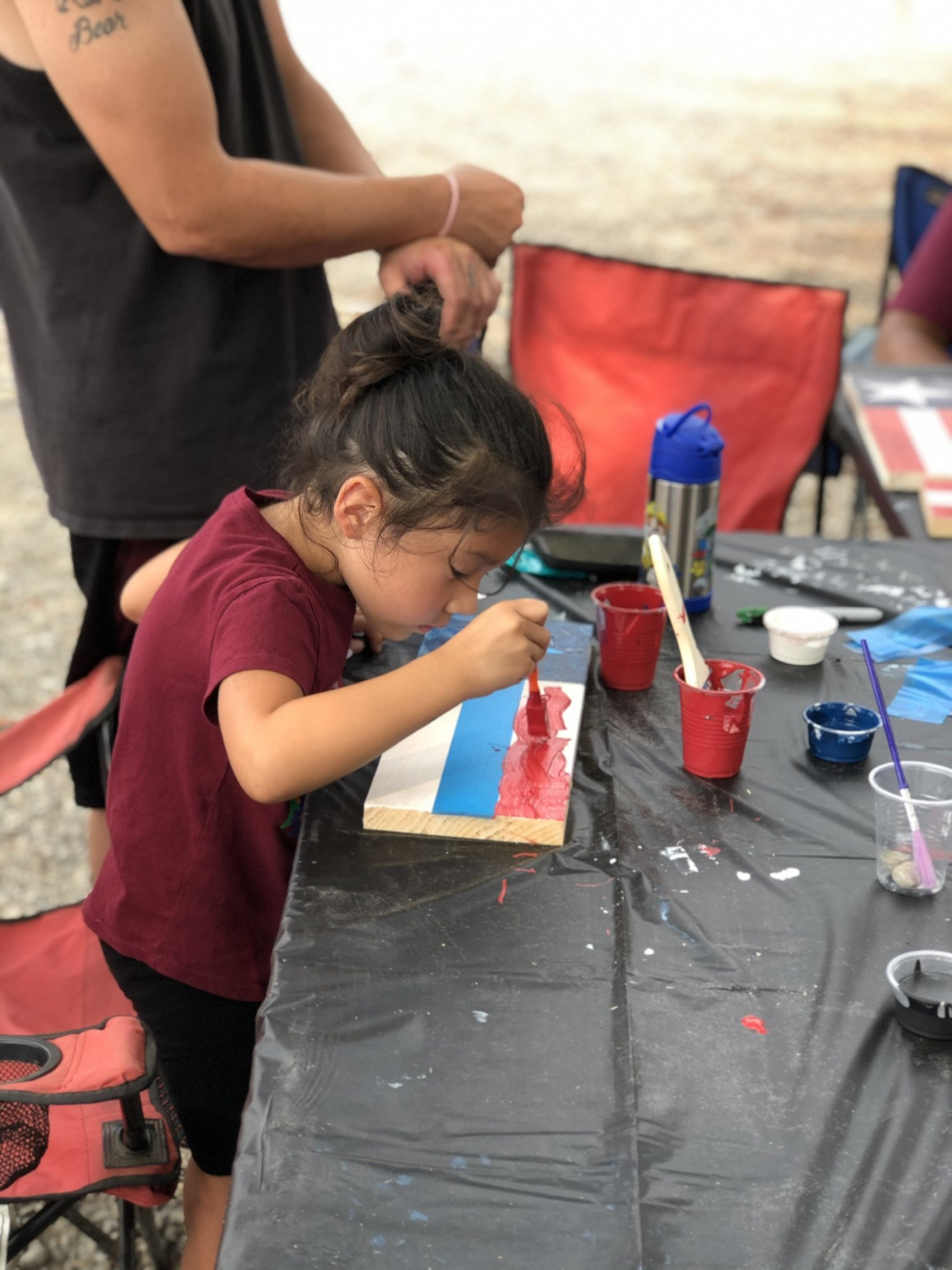 A young girl painting on a wooden plank with red paint, sitting at a table covered with a black plastic tablecloth, with art supplies around her, under an adult's supervision.