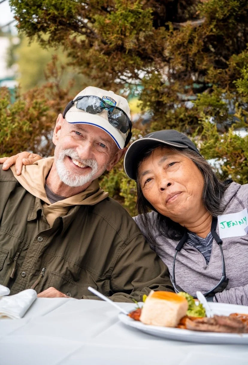 Two smiling people, a man and a woman, sitting at a table outdoors with food, surrounded by trees.