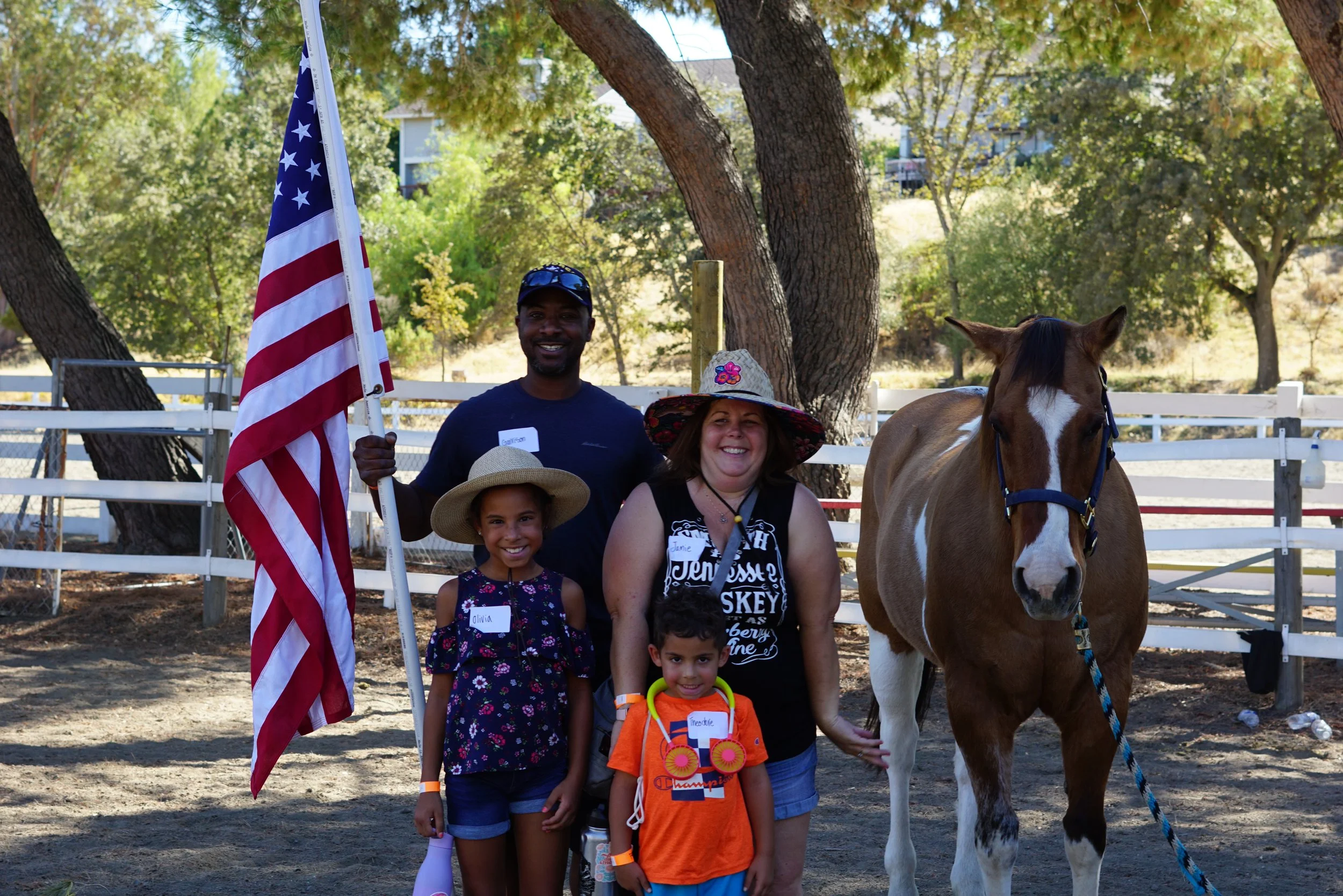 A group of five people, including children and adults, posing outdoors in front of a white fence and trees, with a brown and white horse and an American flag. The group is smiling and wearing casual summer clothing, some with hats, and holding small 