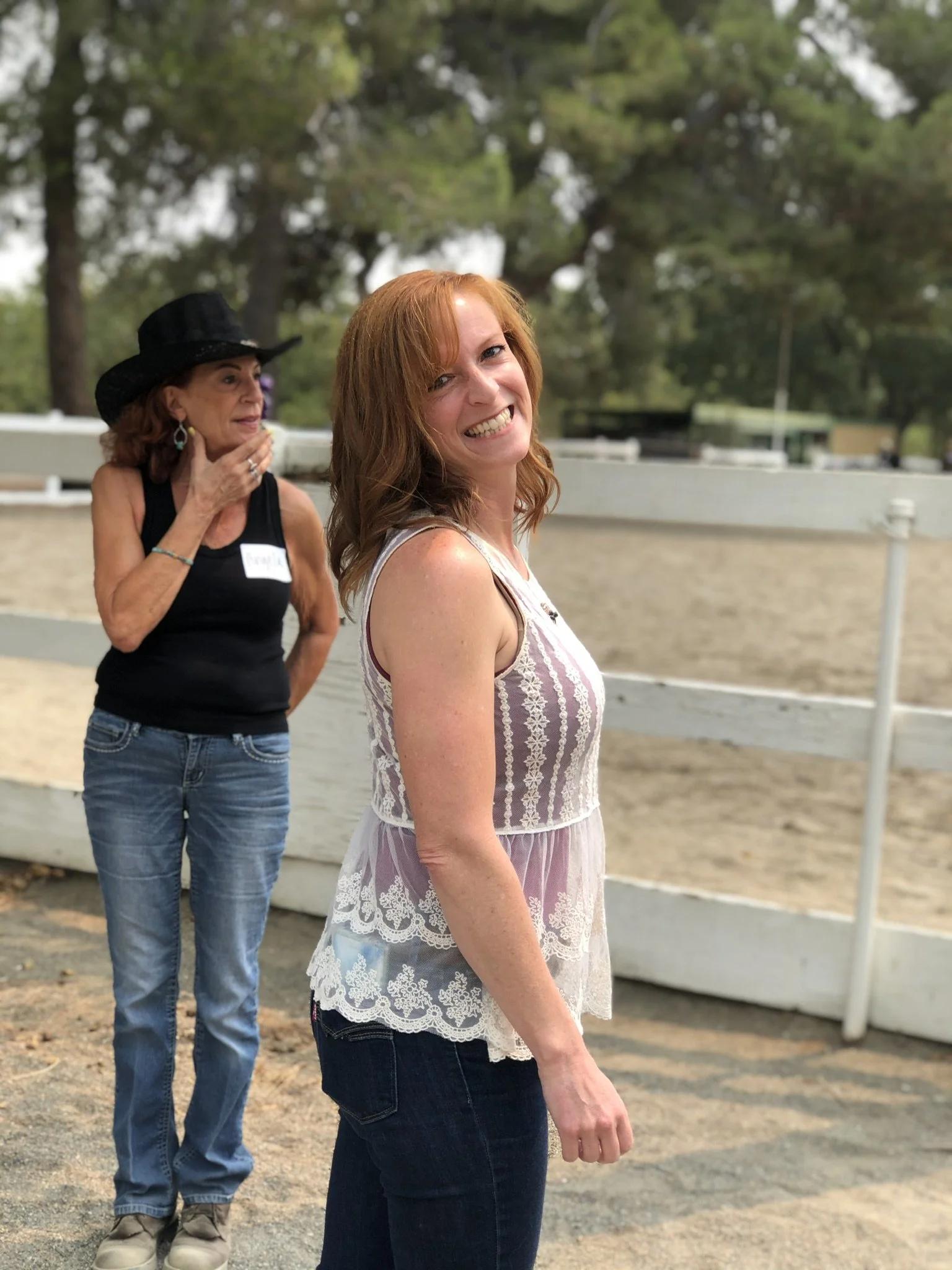 A woman with red hair wearing a light pink and white sleeveless lace top is smiling and walking outdoors, with trees and a fence in the background. An older woman in a black top and cowboy hat stands behind her, touching her chin and looking to the side.