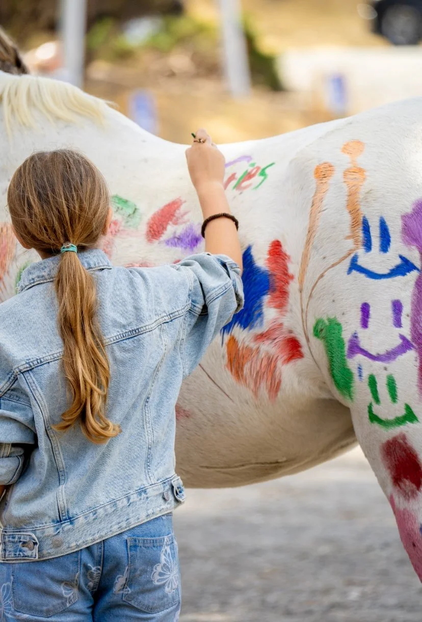 A young girl with long, light brown hair in a ponytail wearing a denim jacket and jeans, painting colorful smiley faces and abstract designs on a white horse with spray paint.