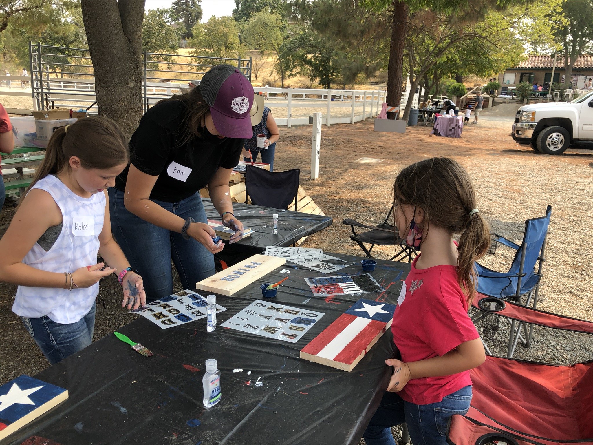 Children and an adult painting patriotic-themed decorations on wooden boards outdoors under trees, with tables, chairs, and a white fence in the background.