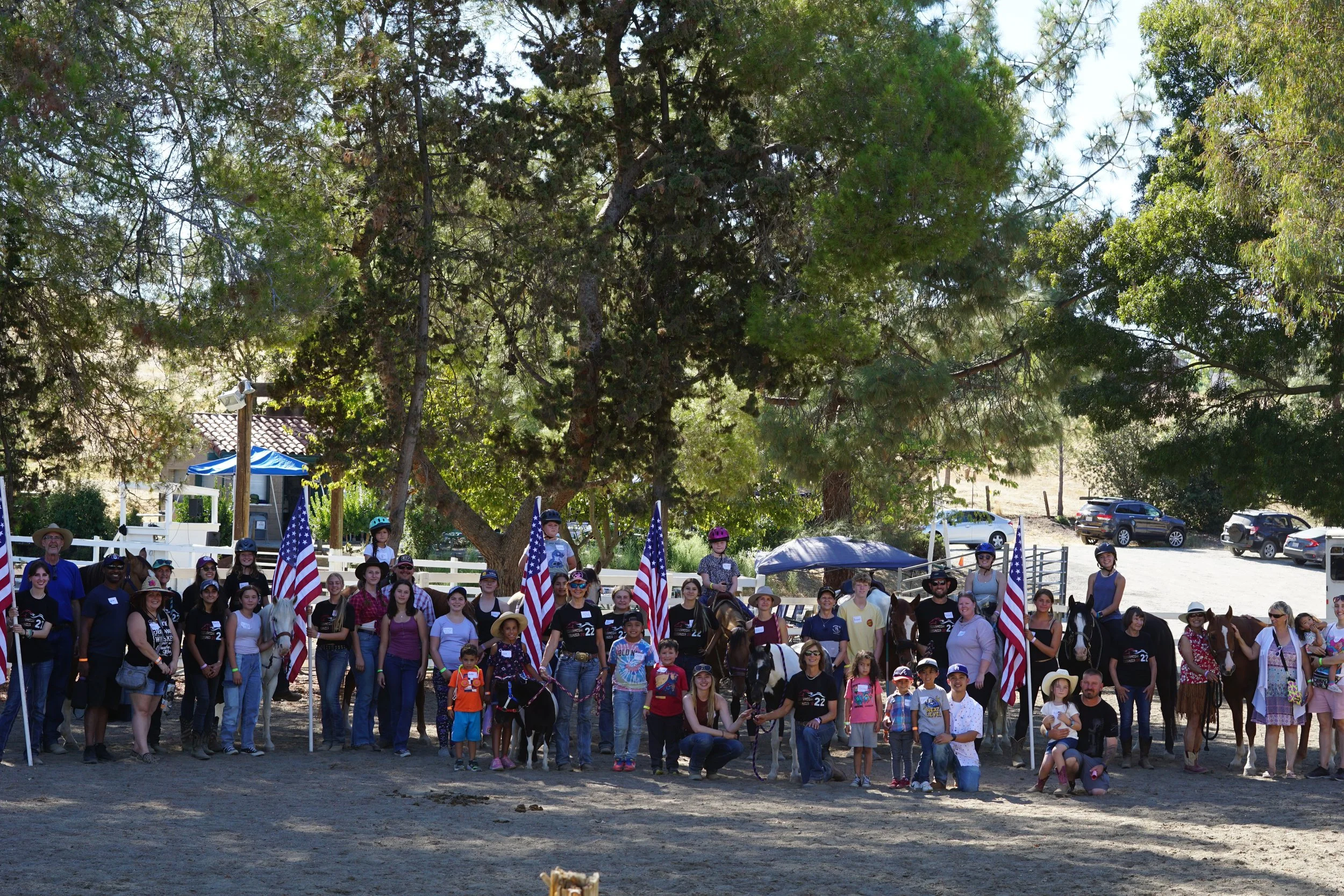 Group photo of people and children gathered outdoors, some on horses, with American flags and trees in the background.