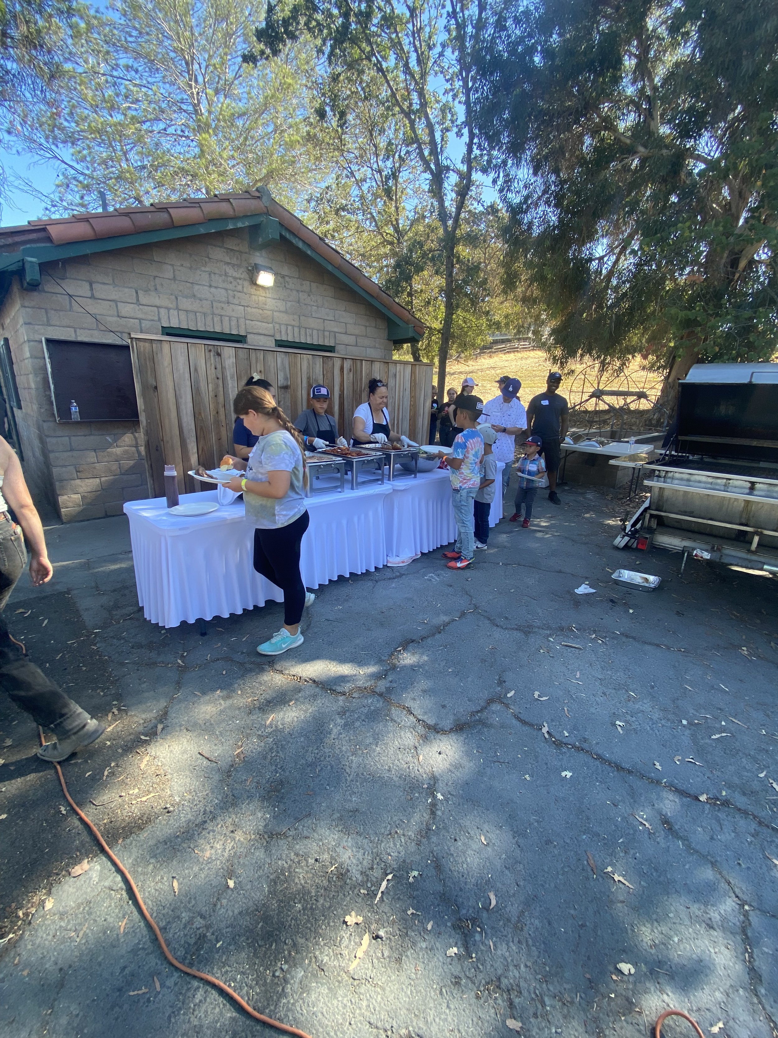 People are gathered outdoors around a table with food, with some serving and others waiting, on what appears to be a sunny day, outside a house with trees in the background.