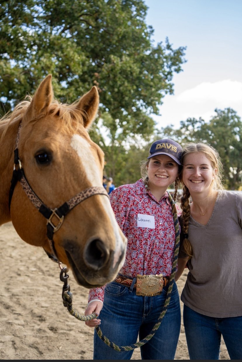 Two young women smiling, one holding a light brown horse's reins, outdoors with trees and blue sky in the background.