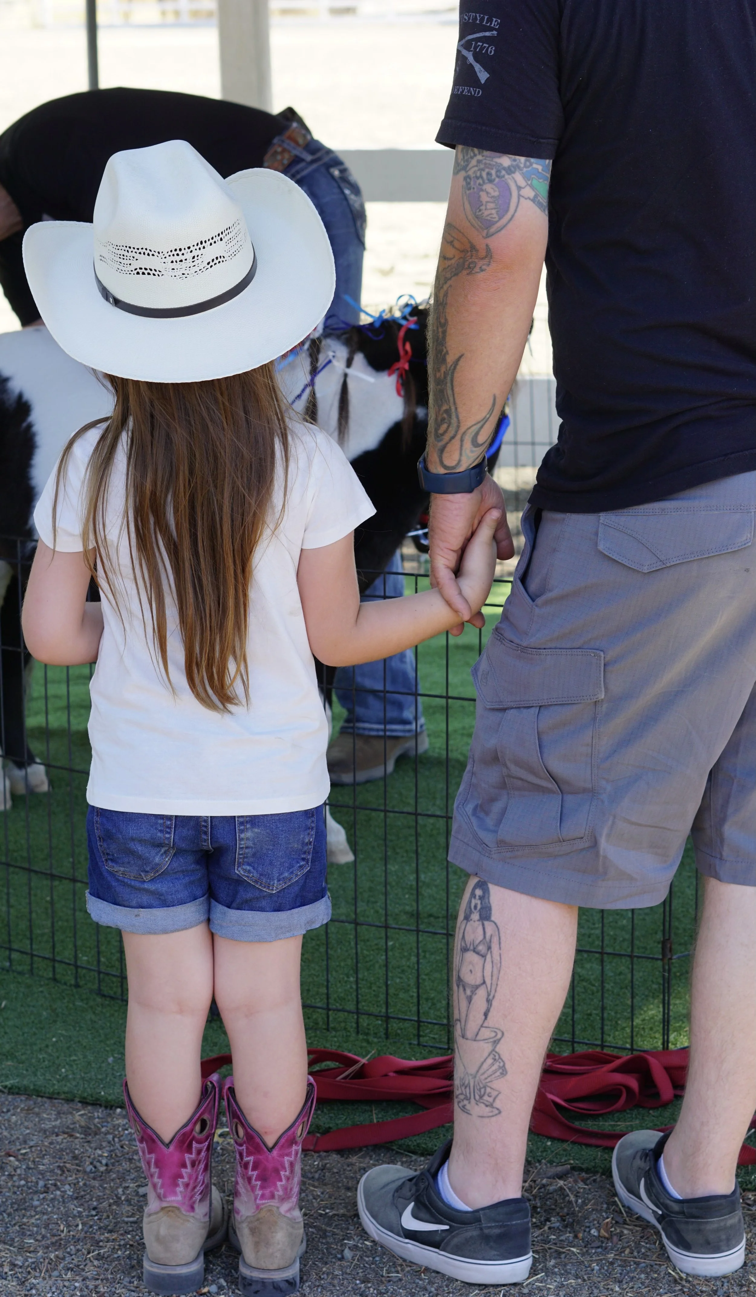 A young girl in denim shorts and cowboy boots holding hands with an adult man with tattoos on his arm and leg, standing at a petting zoo with goats in the background.