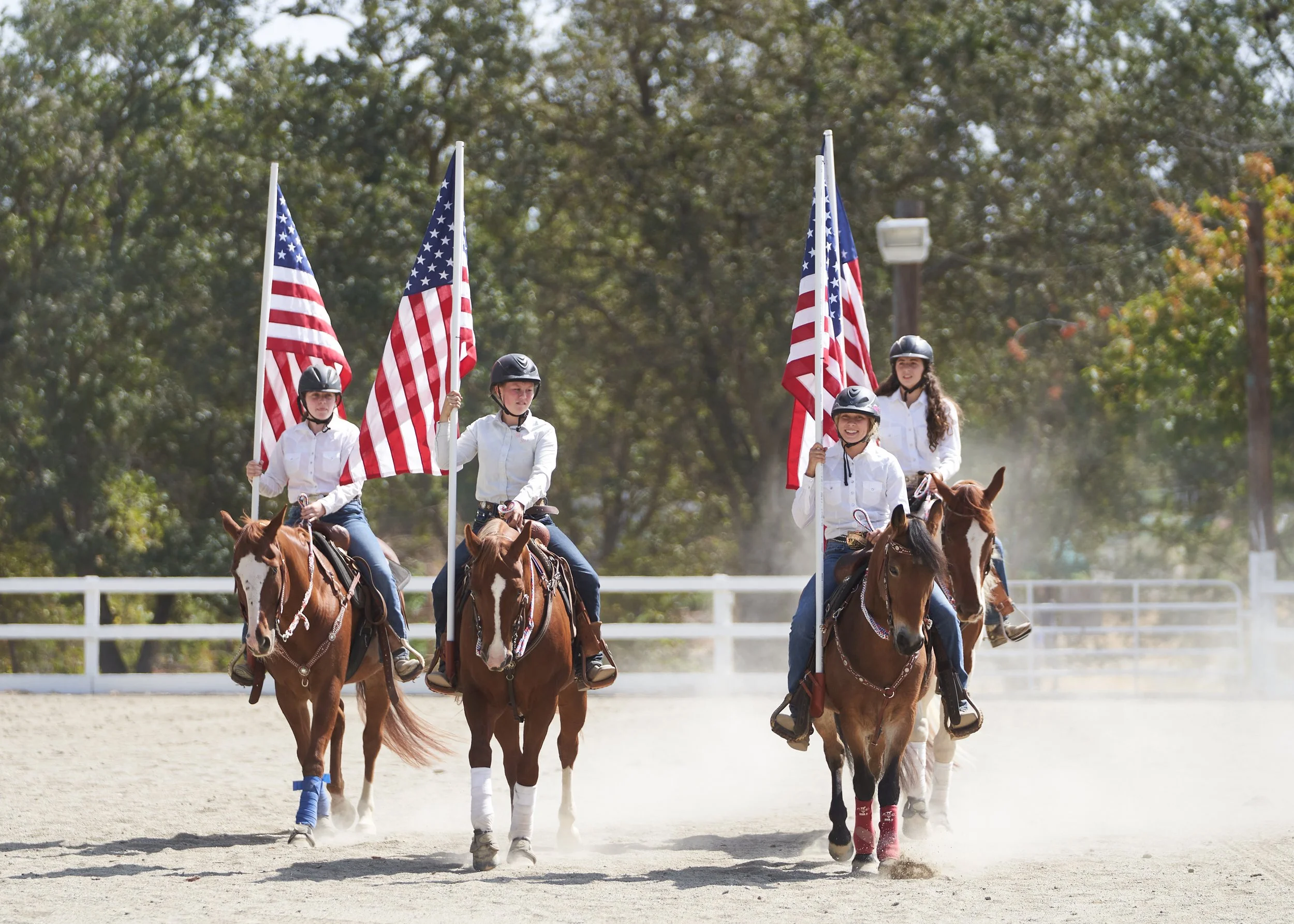 Four young women riding horses and holding American flags outdoors on a sunny day with trees in the background.