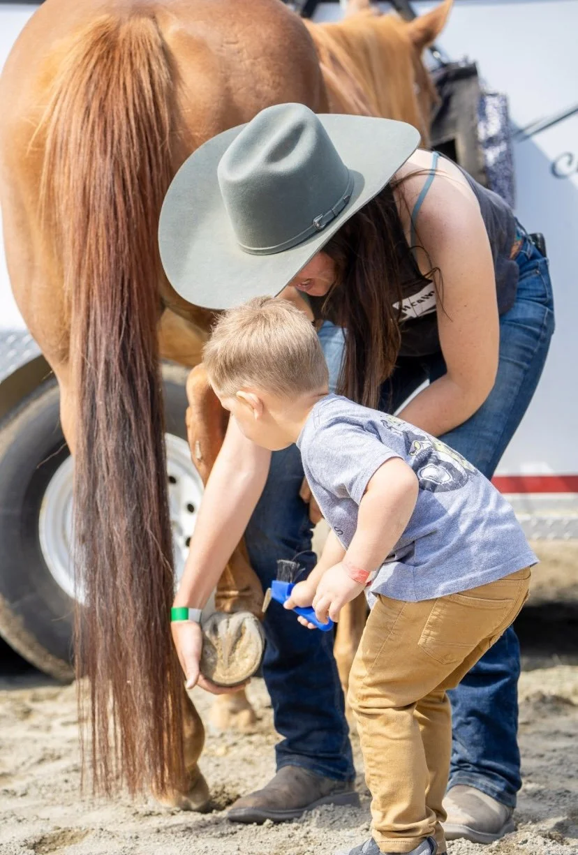 A woman with a large gray hat and blue jeans helping a young boy with a toy shovel groom and clean a horse's hoof at a farm or petting zoo.