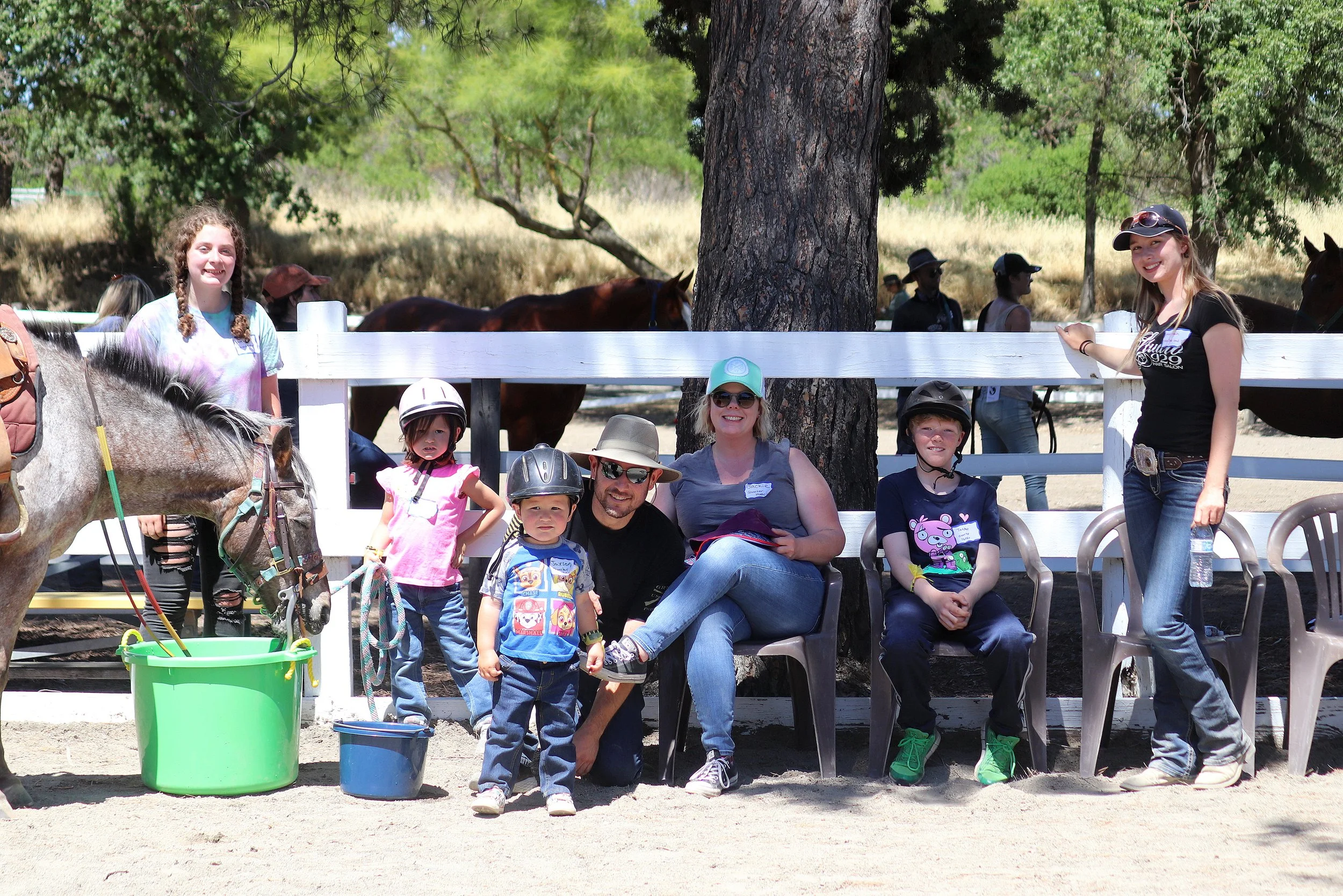 Group of children and adults at a horse riding event. Some children are wearing helmets, and there are horses in the background. A woman is smiling and sitting among the children, with others standing or sitting nearby. The scene is outdoors with tre